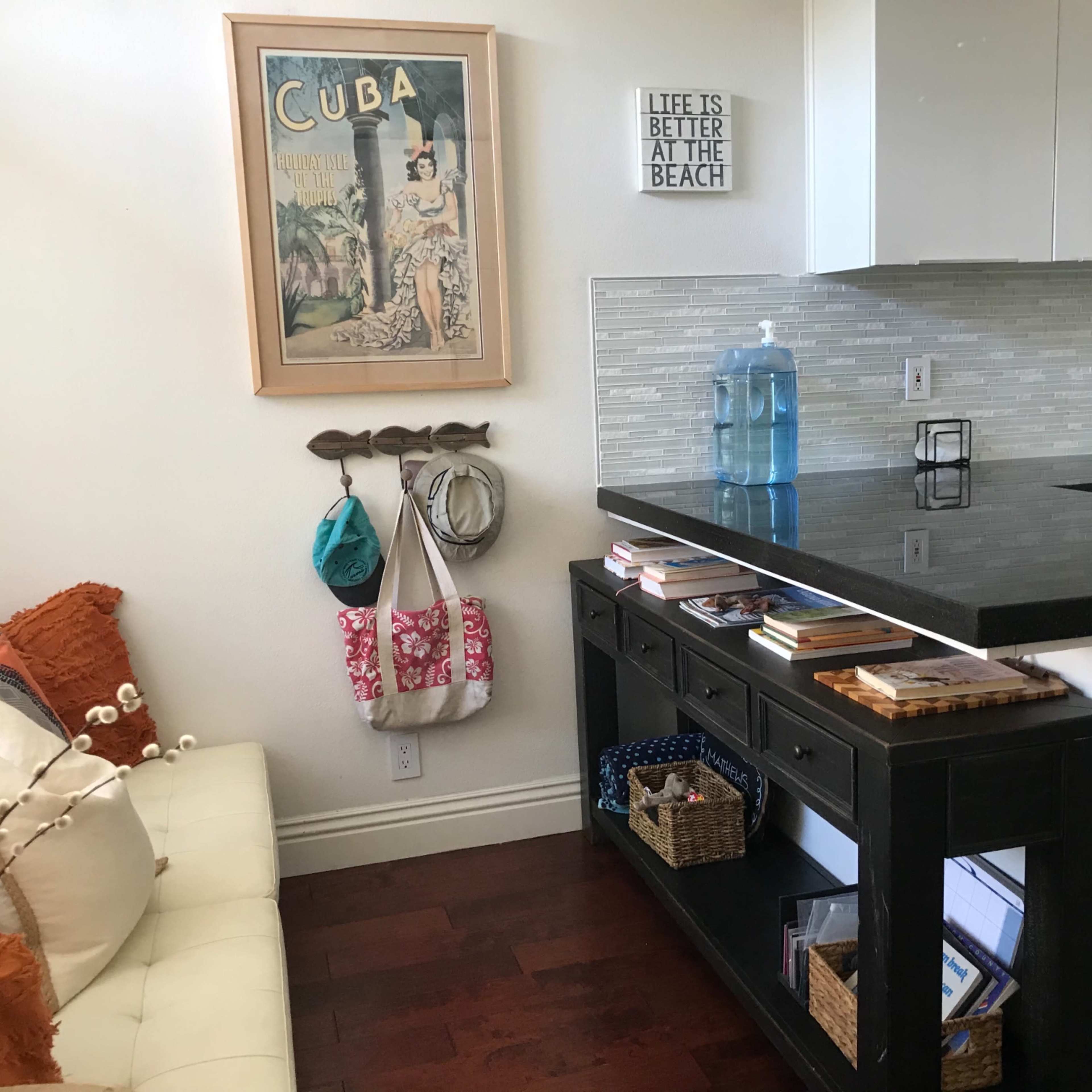 A modern kitchen area with a couch, framed artwork of Cuba on the wall, and a decorative sign that reads "LIFE IS BETTER AT THE BEACH."