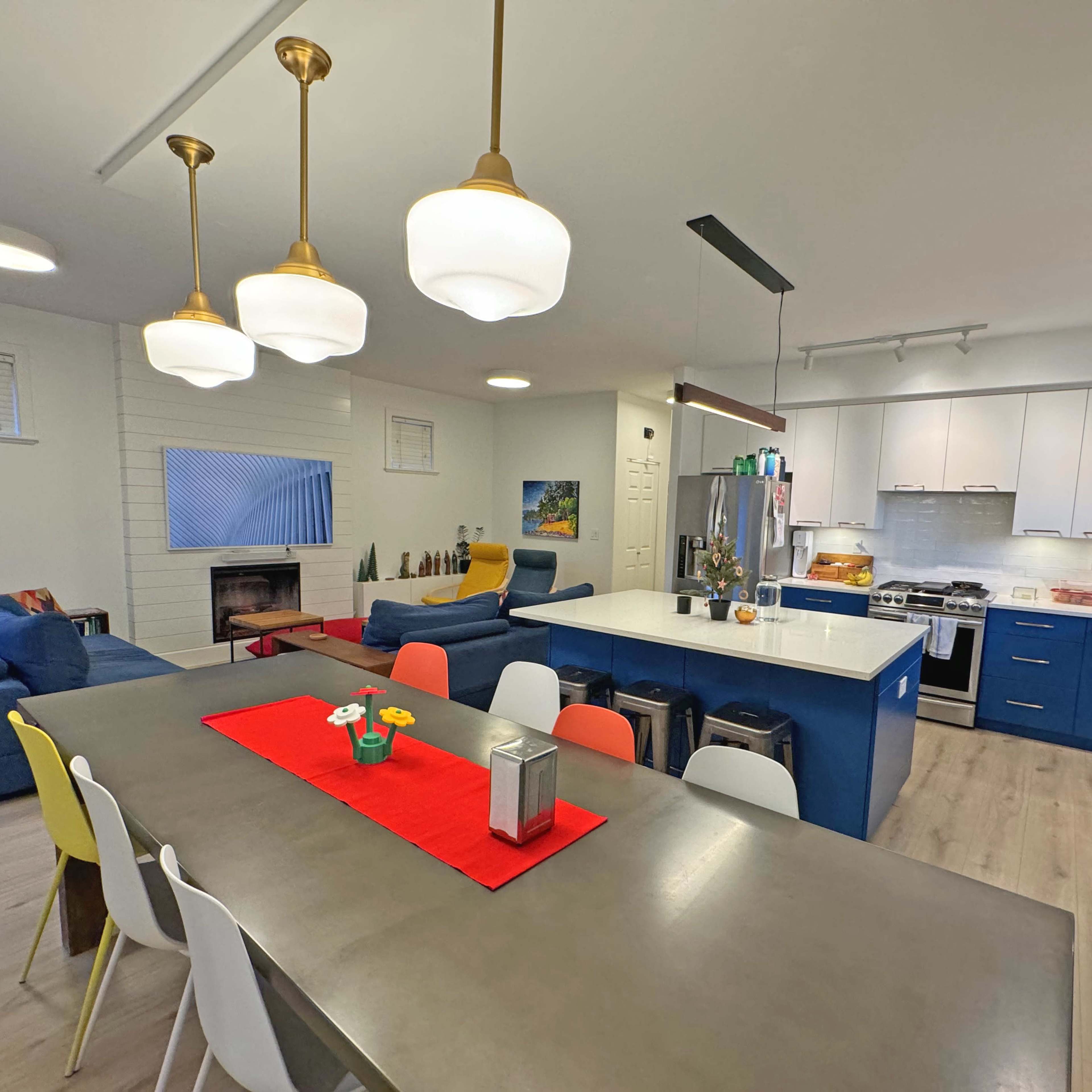 A modern kitchen and dining area with a central table, colorful chairs, and a large island featuring appliances and pendant lighting.