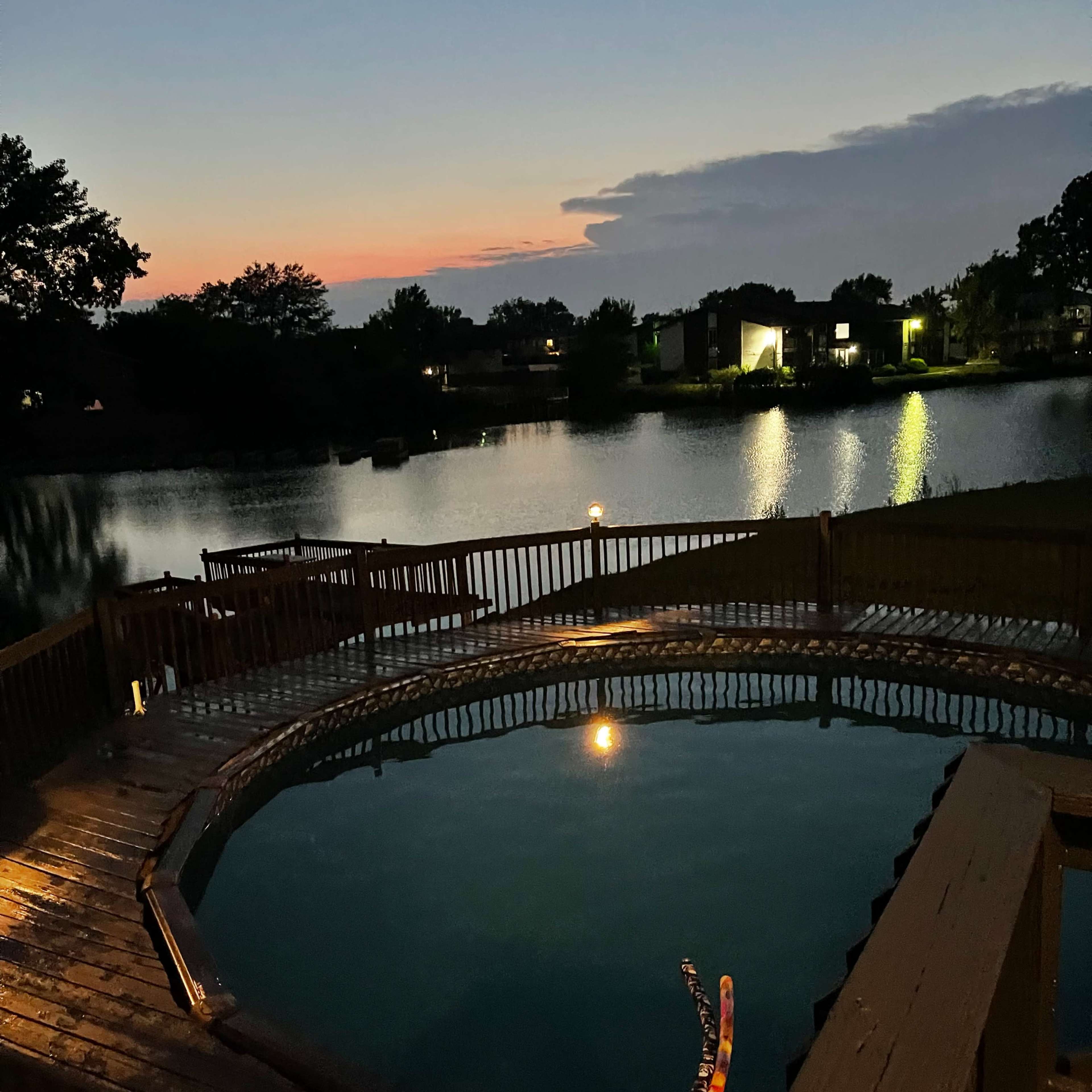 A wooden deck surrounds a circular pool at dusk, reflecting the colors of the sunset over a calm lake.
