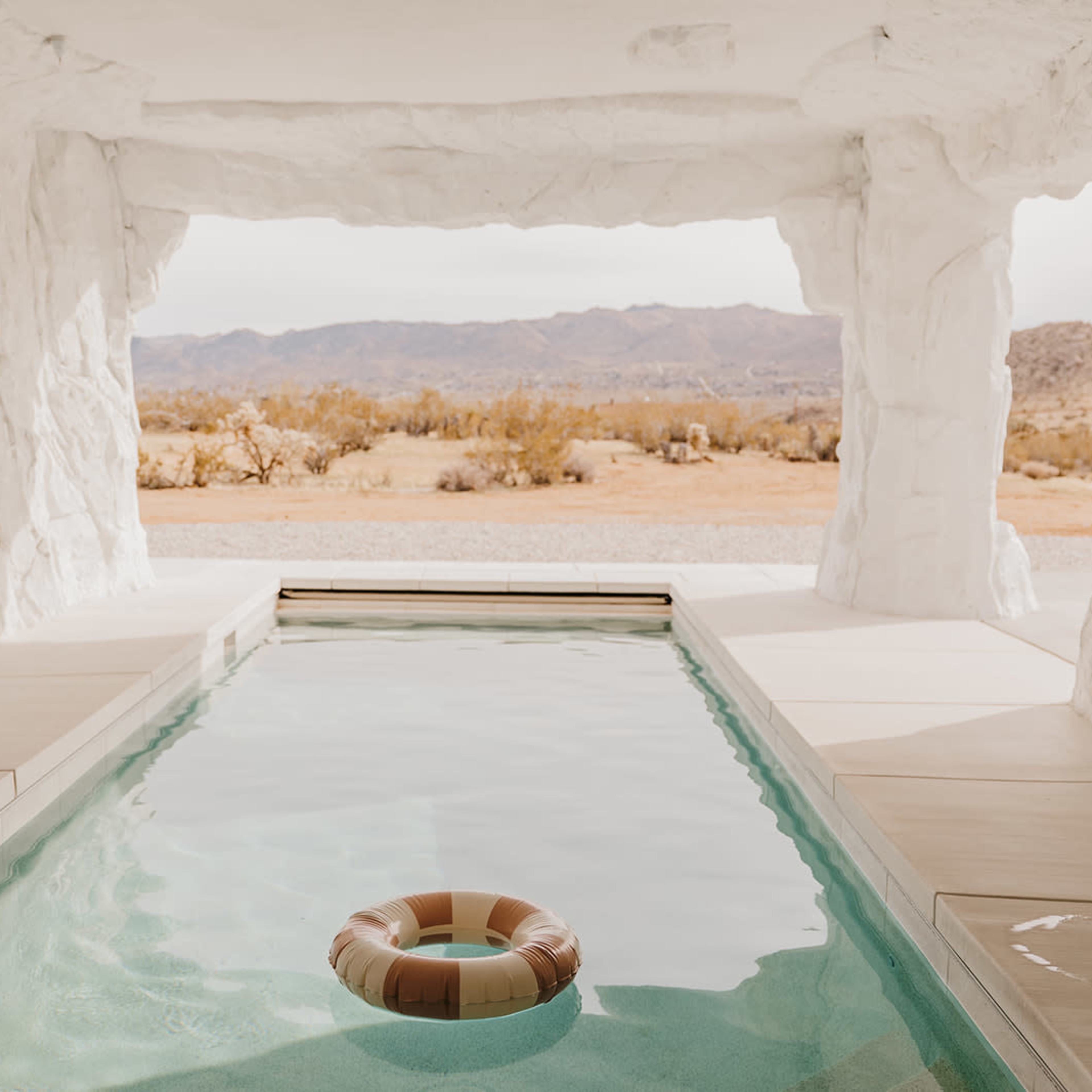 A stone-walled room features a swimming pool with a single inflatable ring, looking out onto a desert landscape.