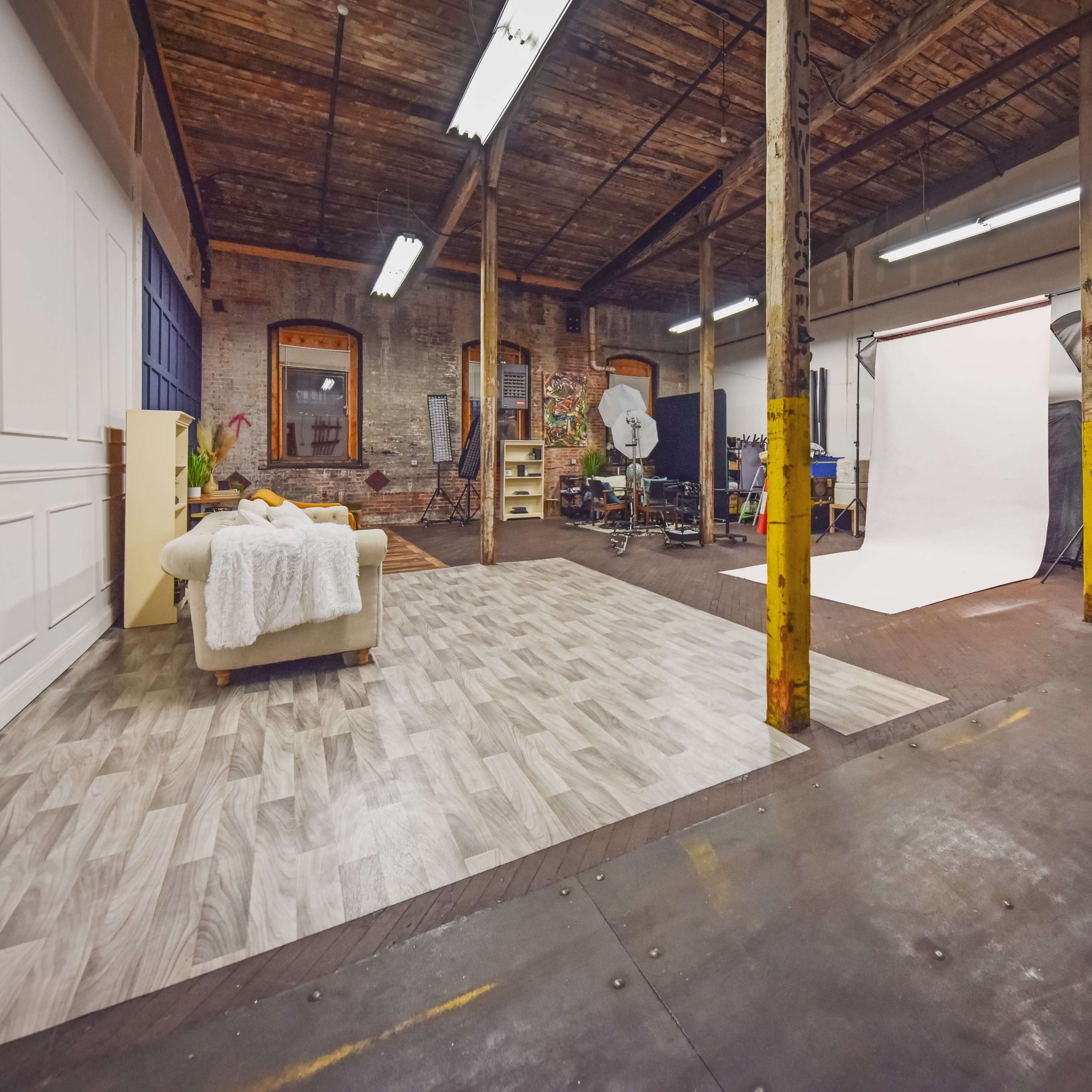 A spacious photography studio featuring a partially furnished area with a couch, a backdrop setup, and various equipment arranged along the walls.