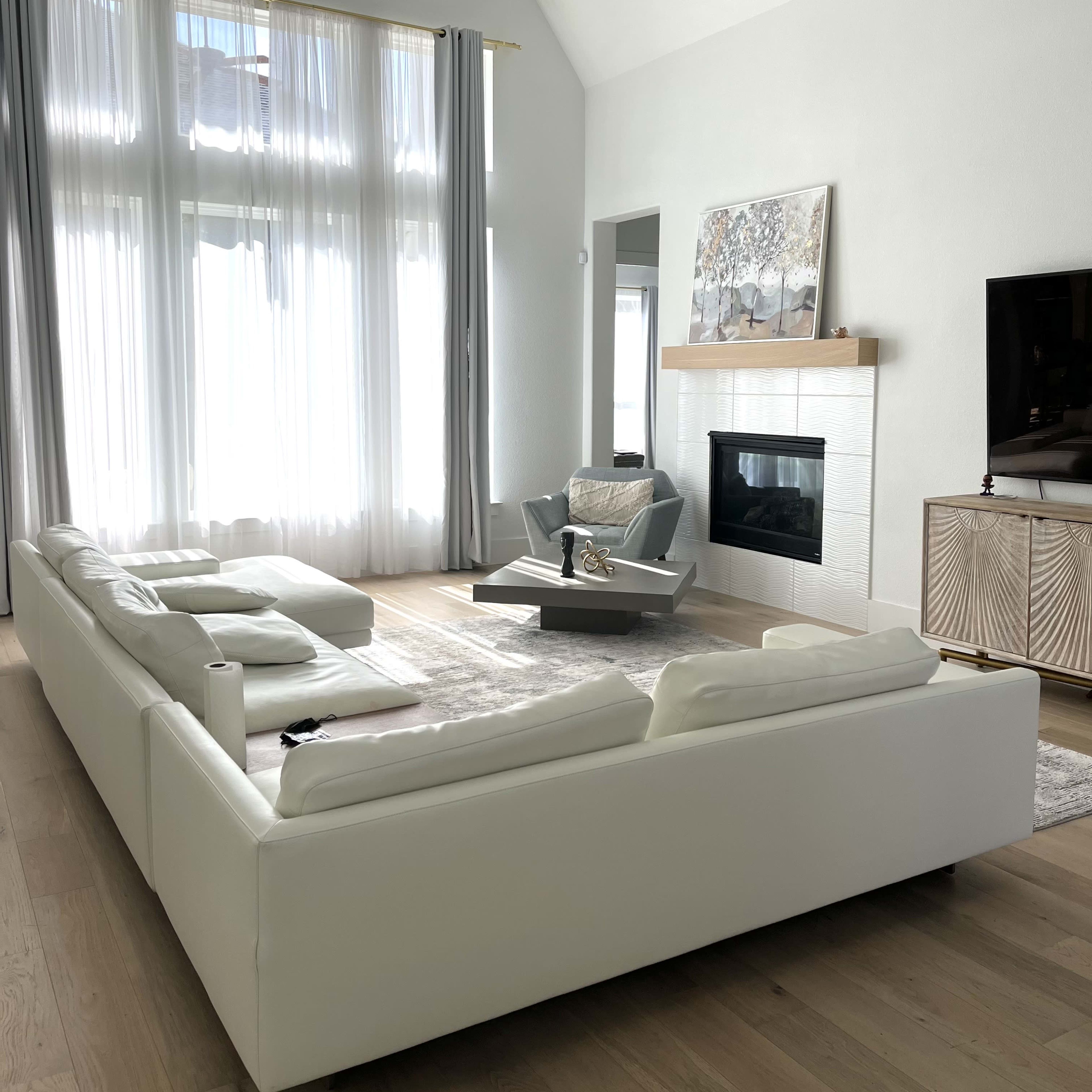 A modern living room with a white sectional sofa, a fireplace, a large window with sheer curtains, and a minimalist coffee table.