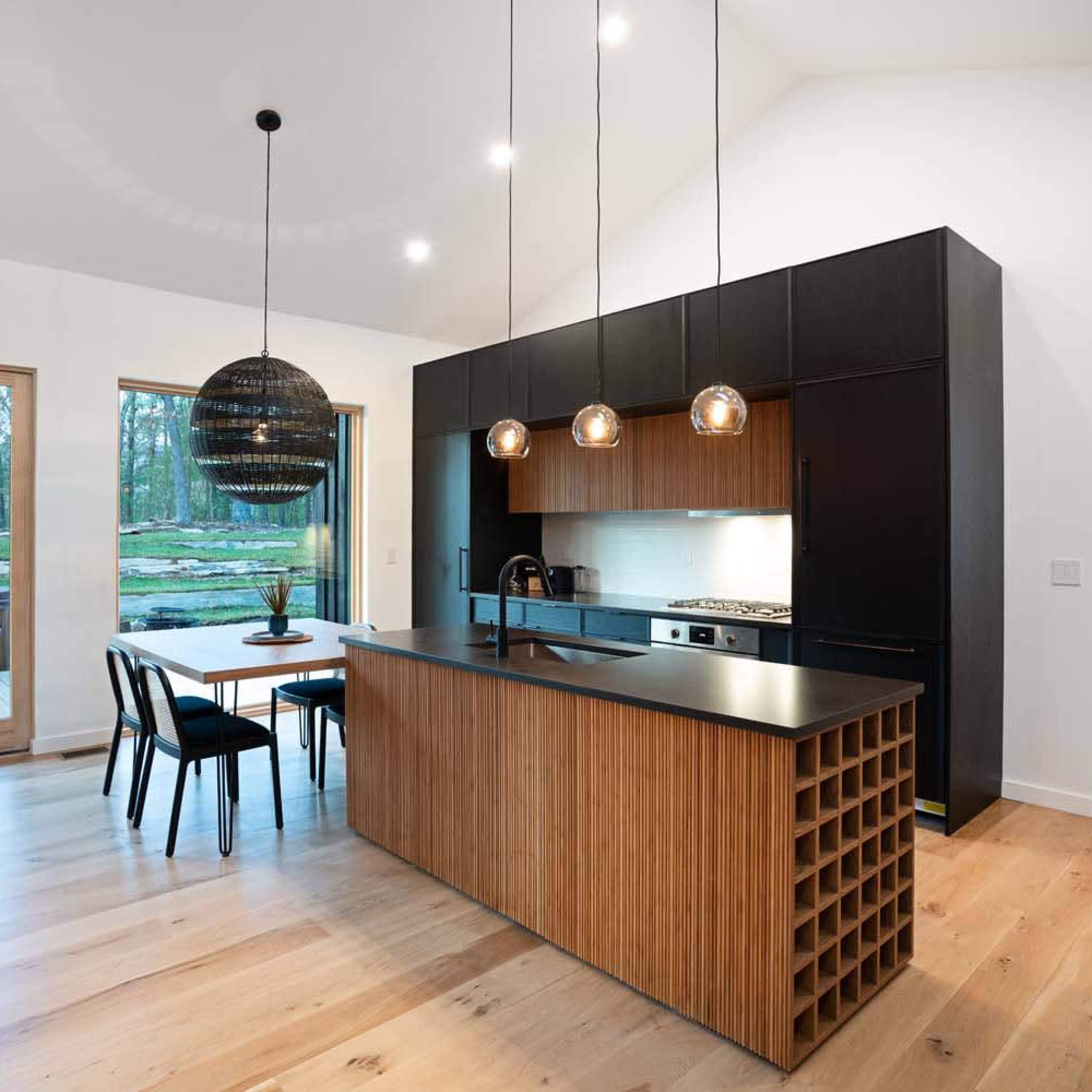 A modern kitchen with a black island, wooden cabinetry, and large windows overlooking a wooded area.