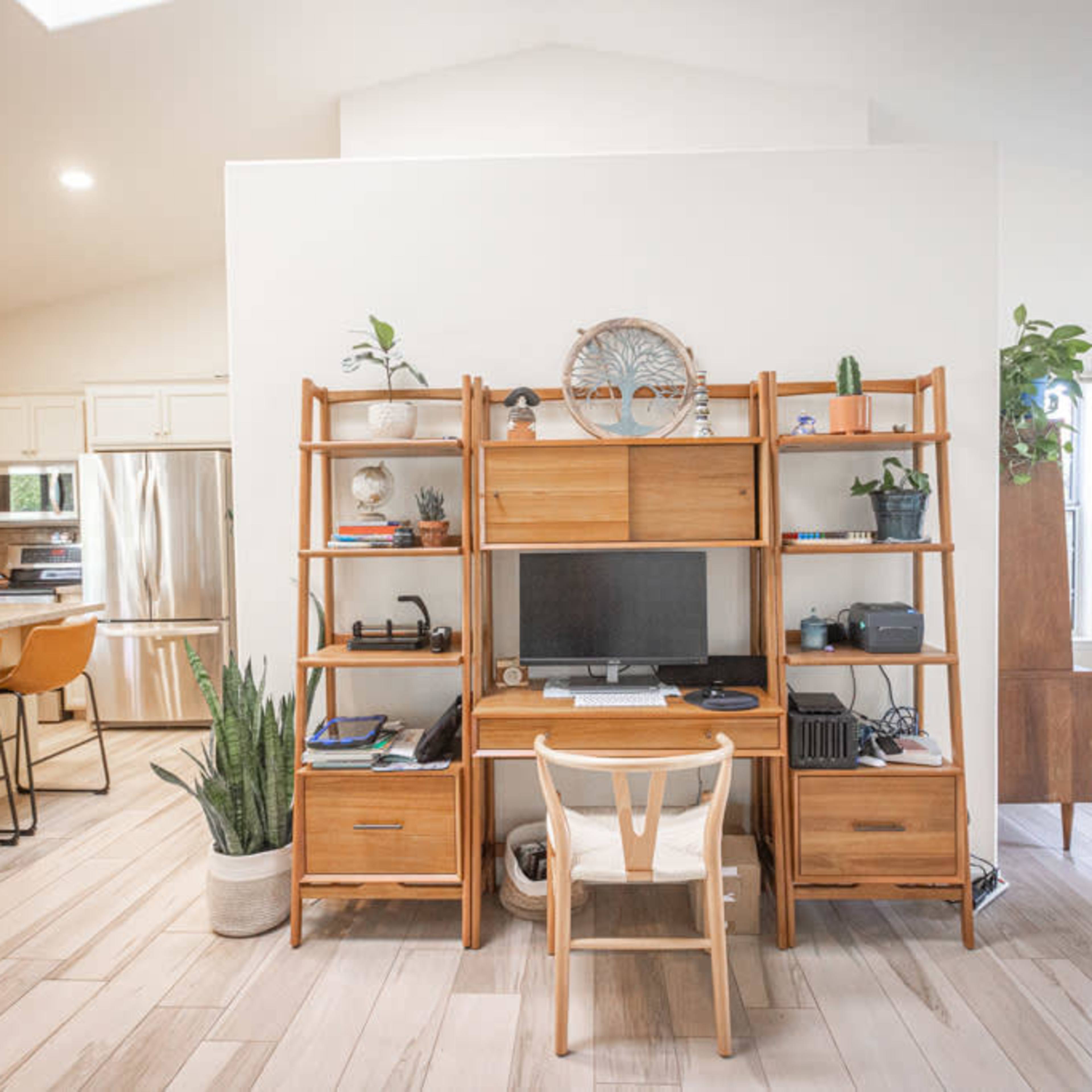 A modern living space with a wooden desk setup against a light wall, featuring a computer and shelves lined with plants and decorative items.