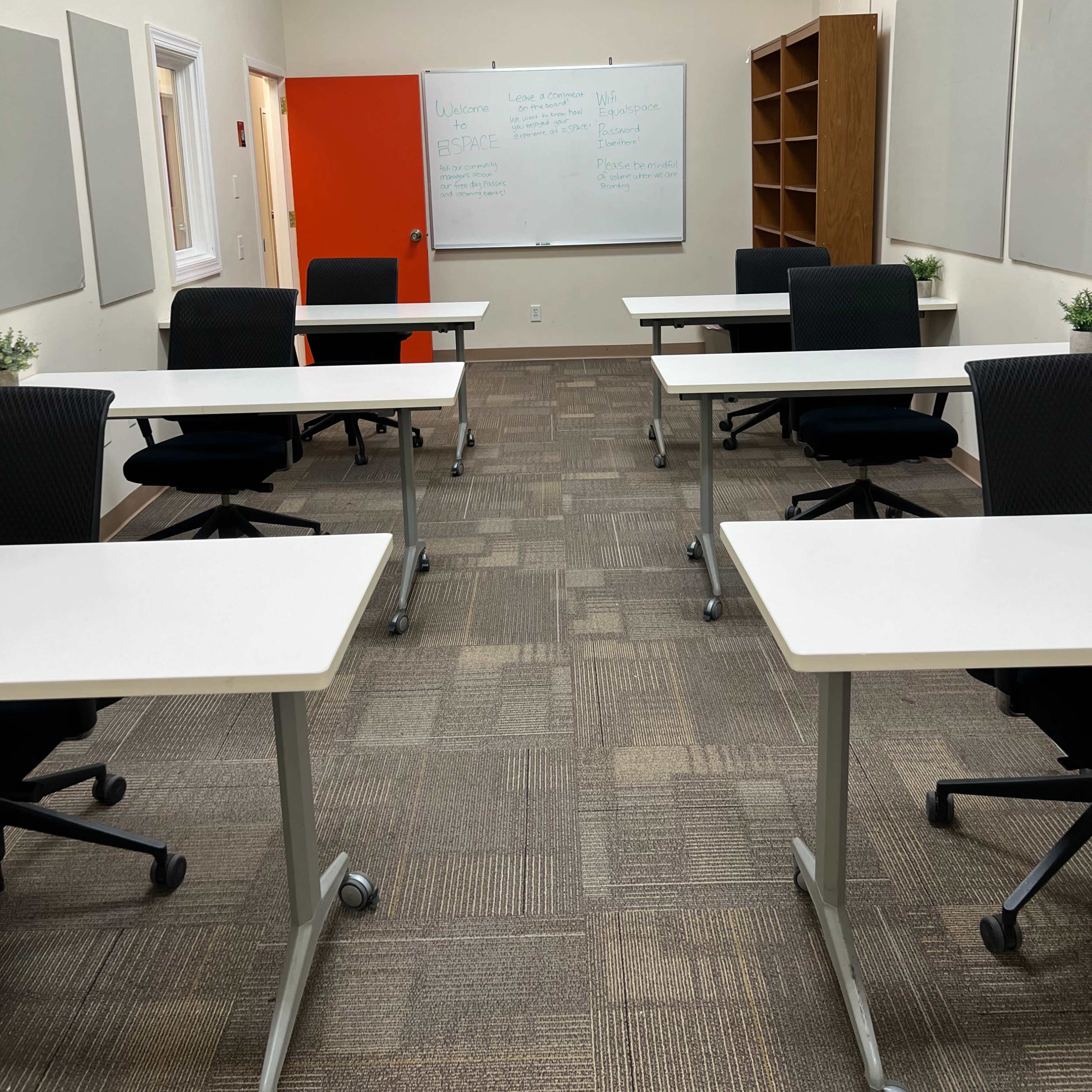 A classroom with several white tables and black chairs arranged around the room, featuring a whiteboard and a red door.