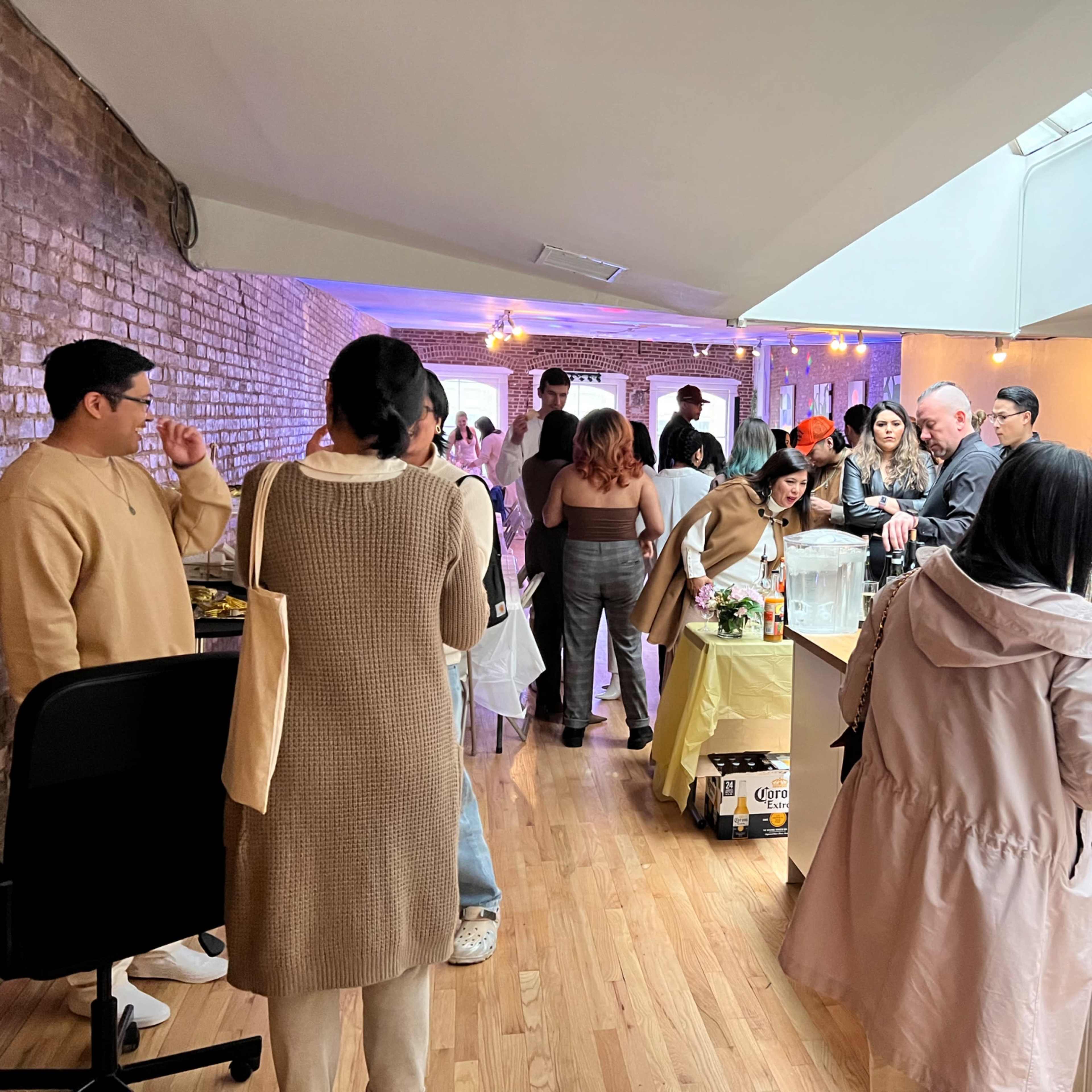A group of people gather in a spacious kitchen area with food and drinks while others socialize in a brightly lit room.
