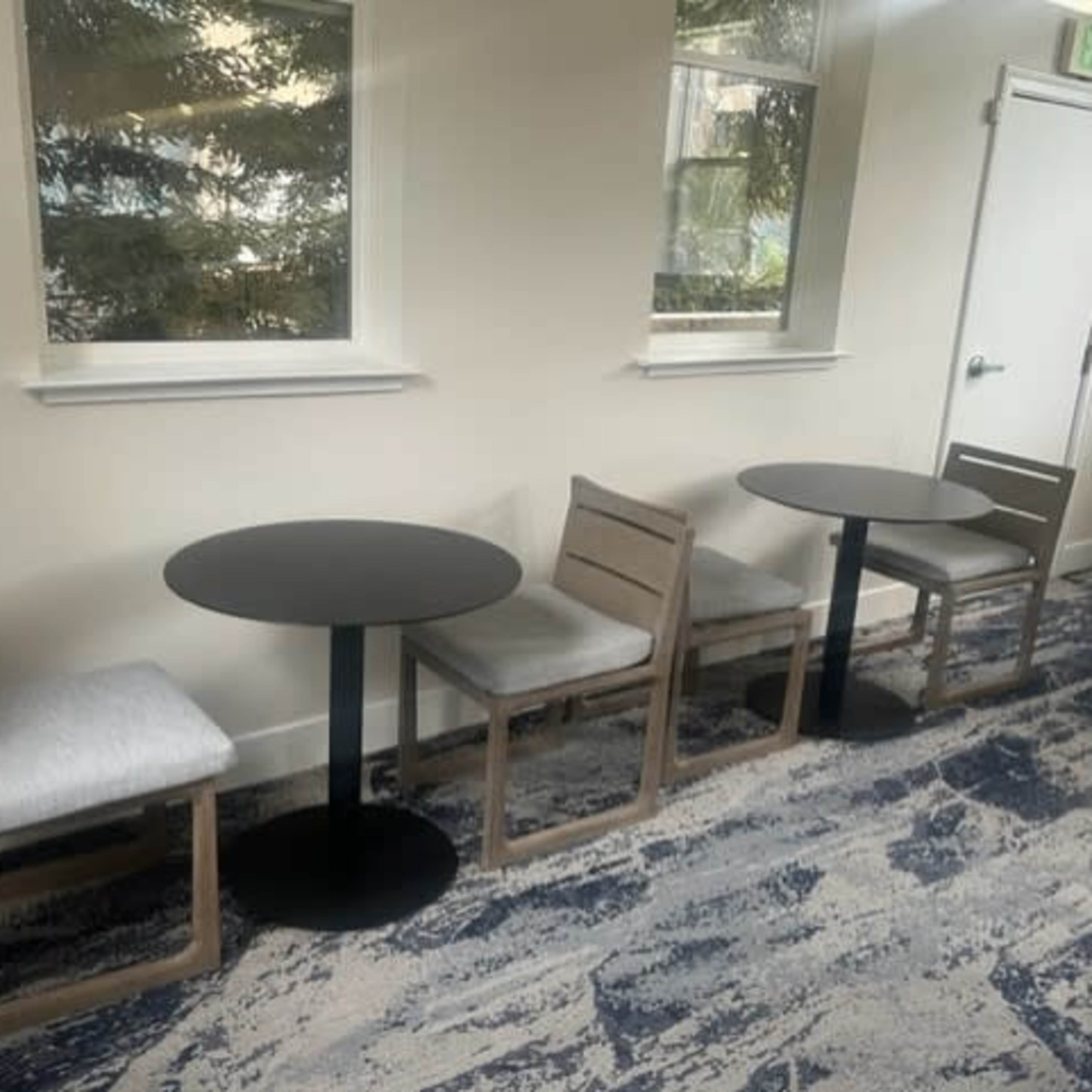 A small area with two round tables and four chairs, situated on a patterned carpet beneath large windows.