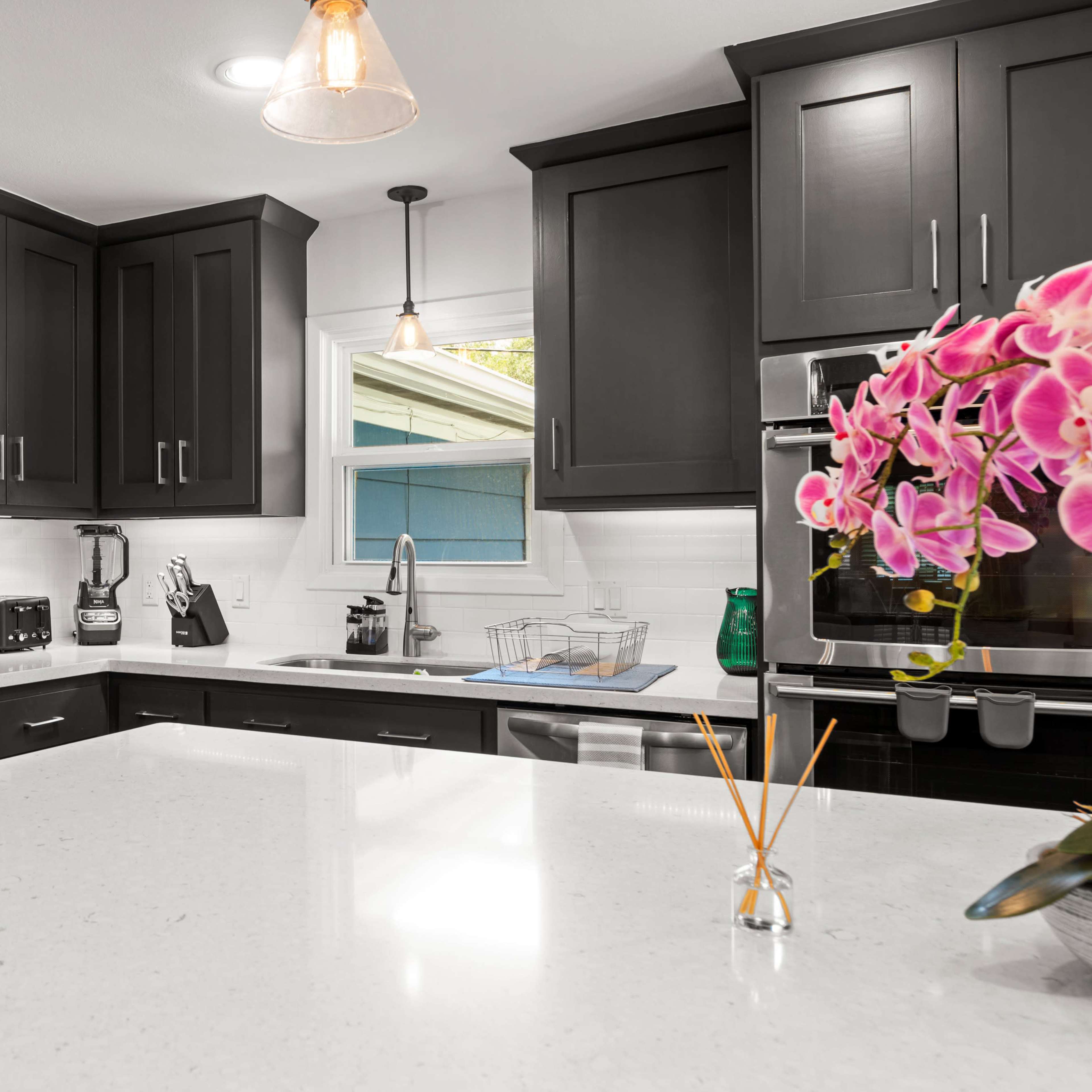 A modern kitchen features dark cabinetry, a large white countertop, and stainless steel appliances, with a decorative orchid on the counter.