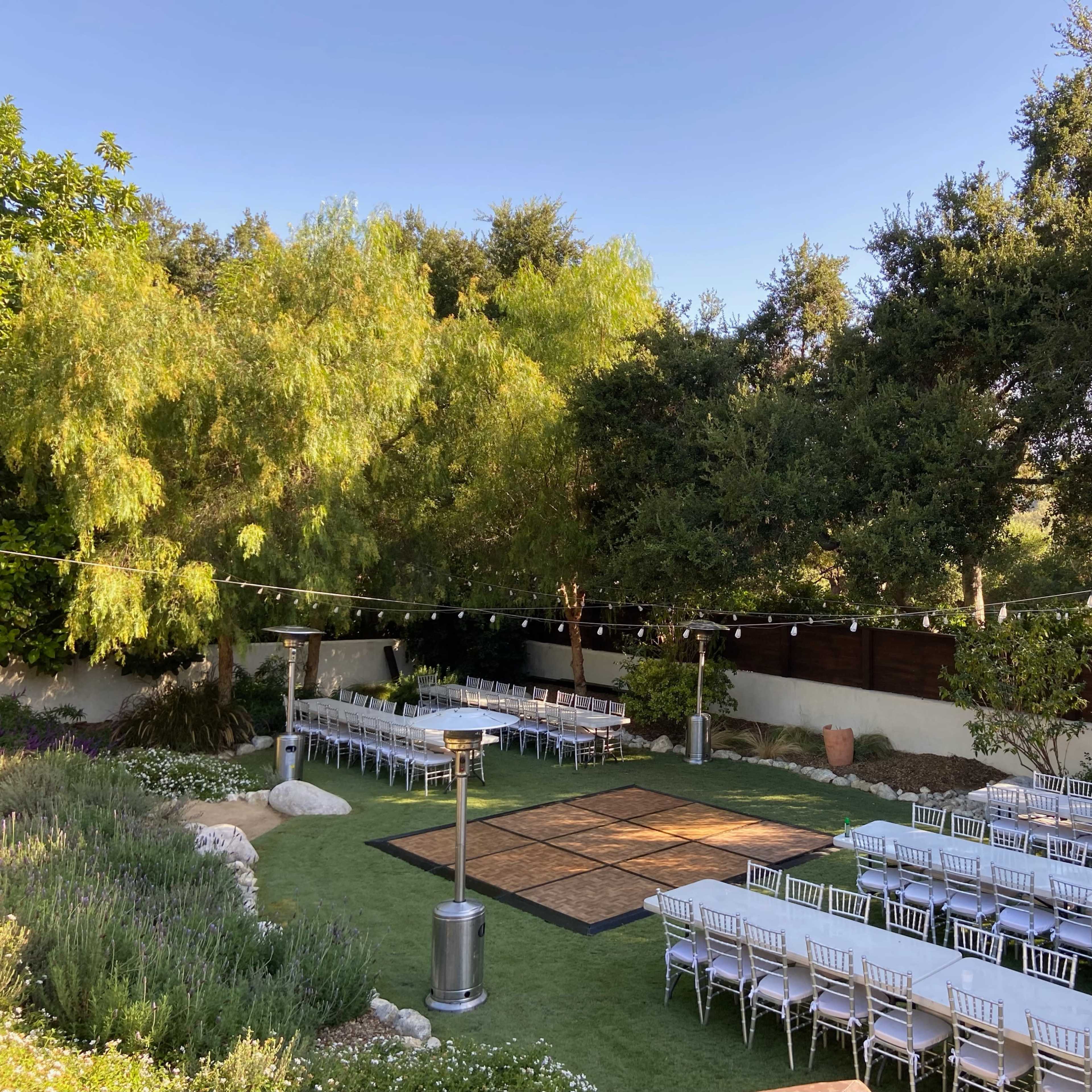 An outdoor event space with neatly arranged rows of tables and chairs set around a dance floor, surrounded by greenery and decorative lights.