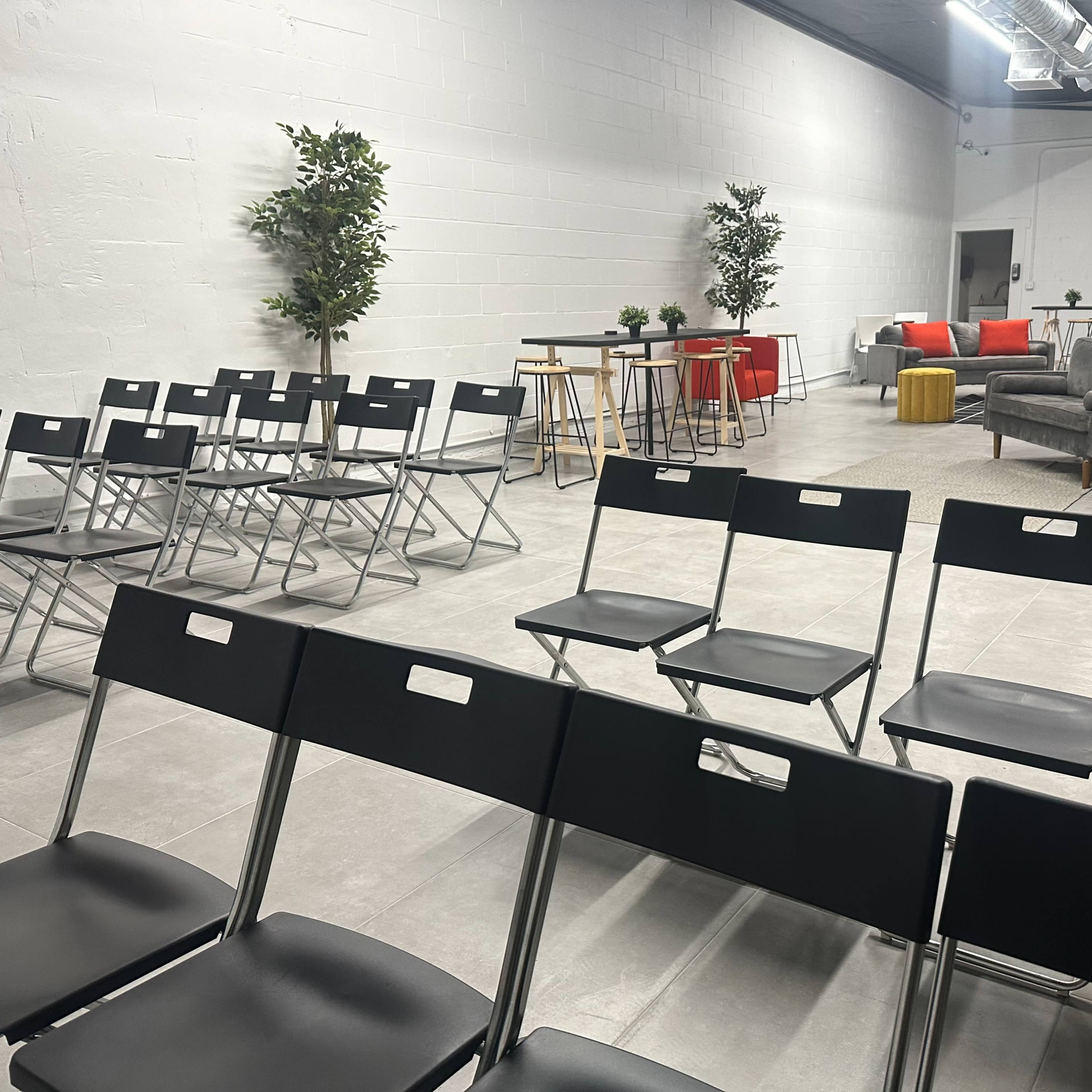 A spacious, modern room arranged with rows of black folding chairs facing a seating area with a couch and tables.