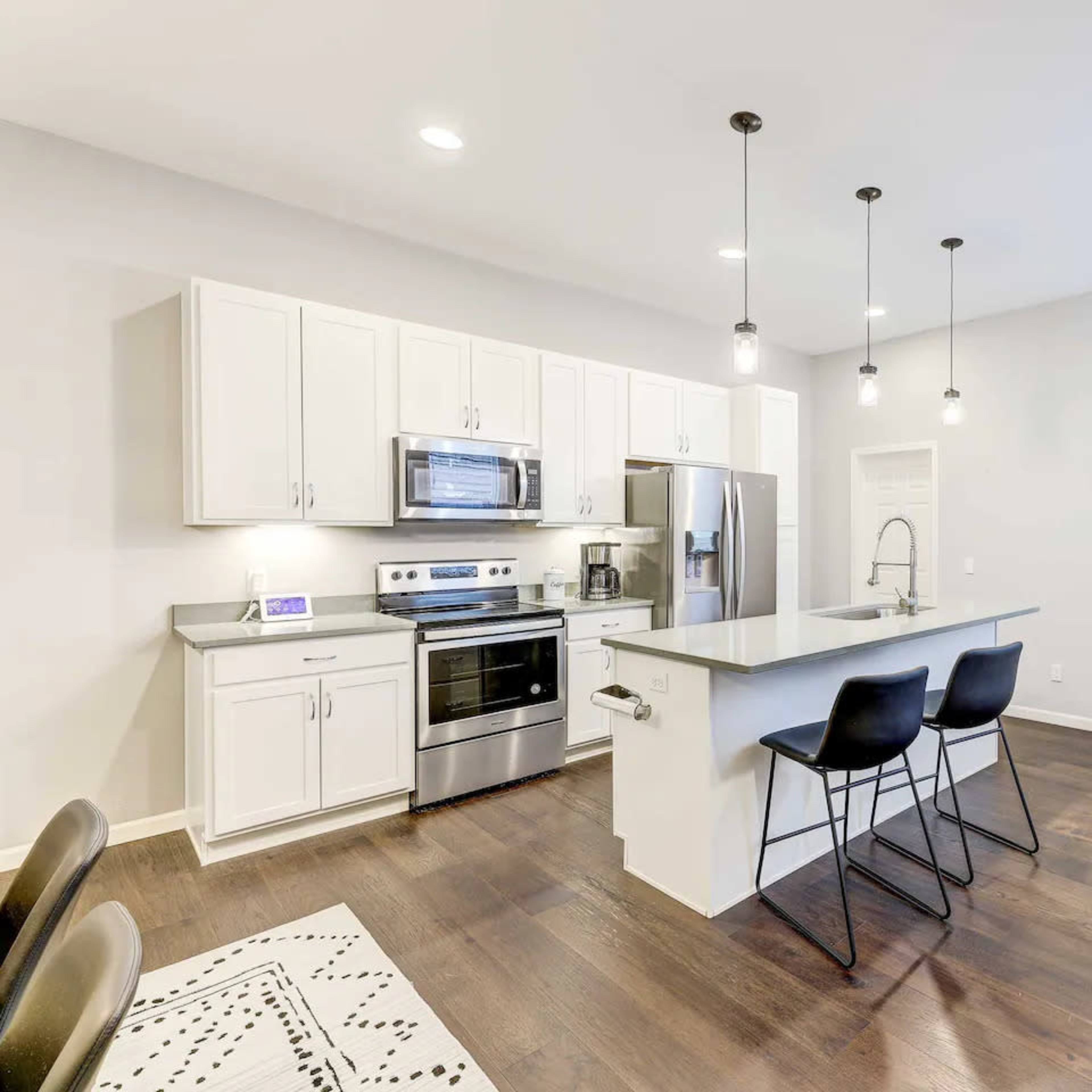 A modern kitchen with white cabinets, stainless steel appliances, and a central island with three black bar stools, leading into a well-lit living area.