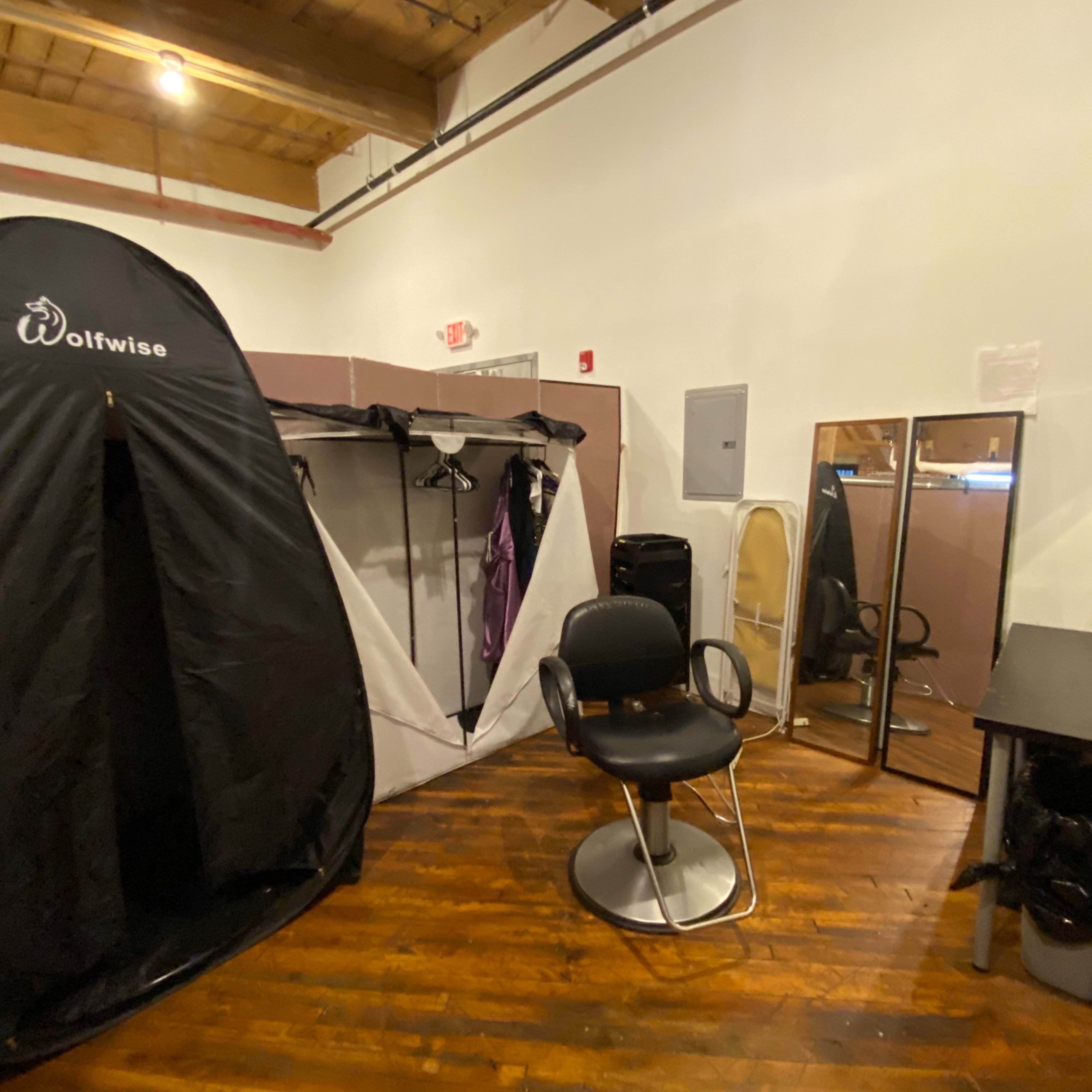 A small room featuring a black privacy tent, a swivel chair, a large mirror, and a table, all set against wooden flooring.