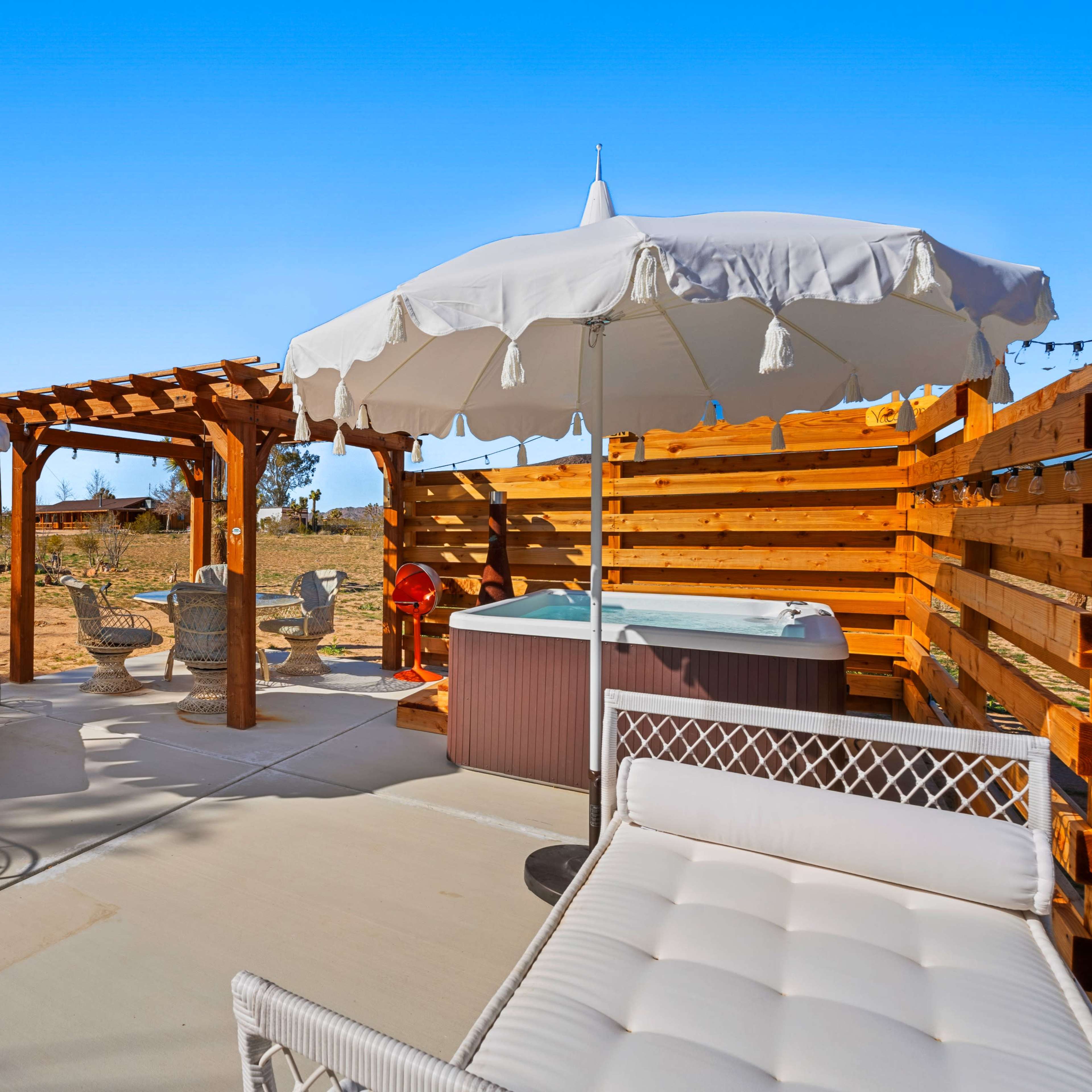 A patio area with a hot tub surrounded by white and pink umbrellas, a wooden pergola, and outdoor seating in a desert landscape.