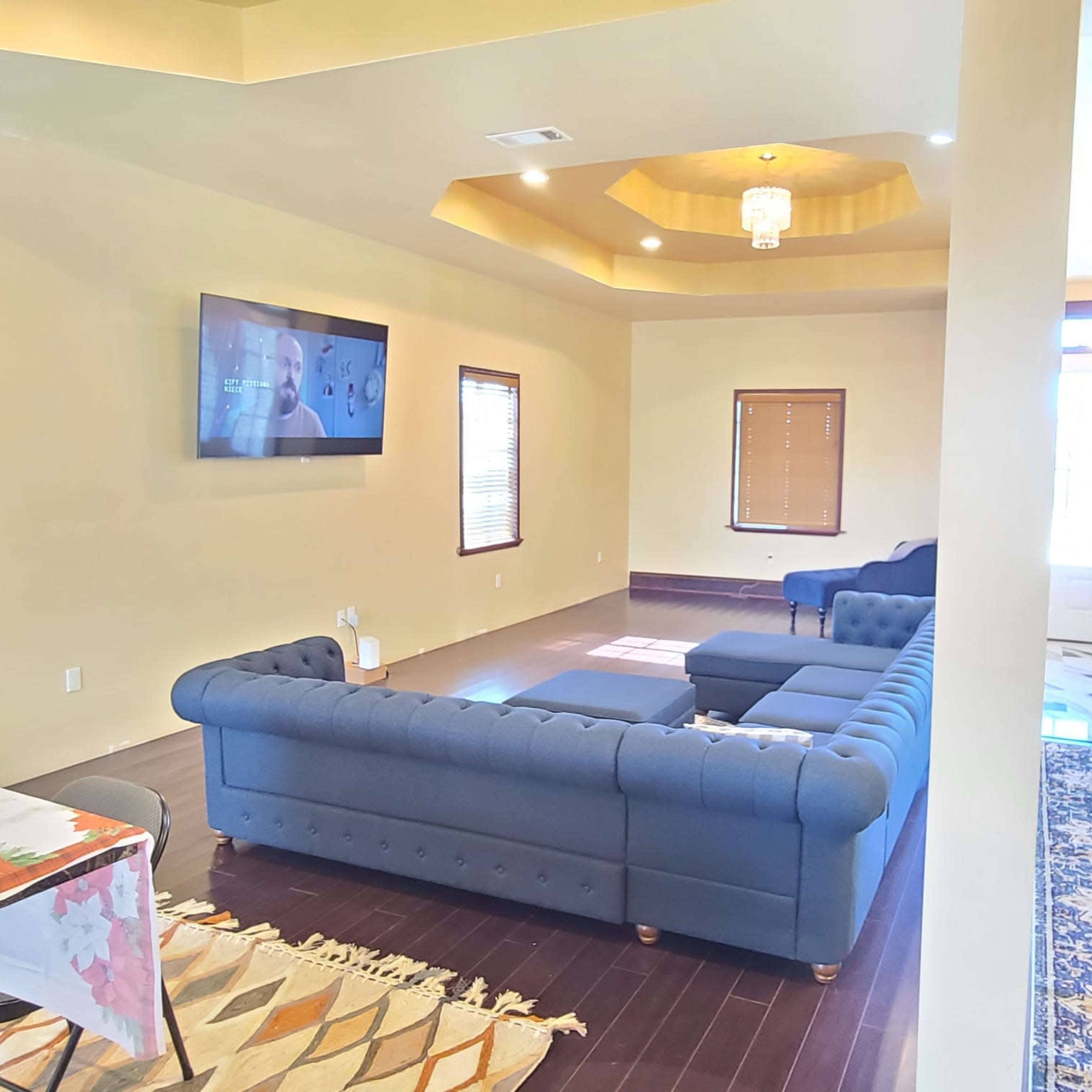 A spacious living room featuring a large blue sectional couch, a television mounted on the wall, and natural light coming through several windows.