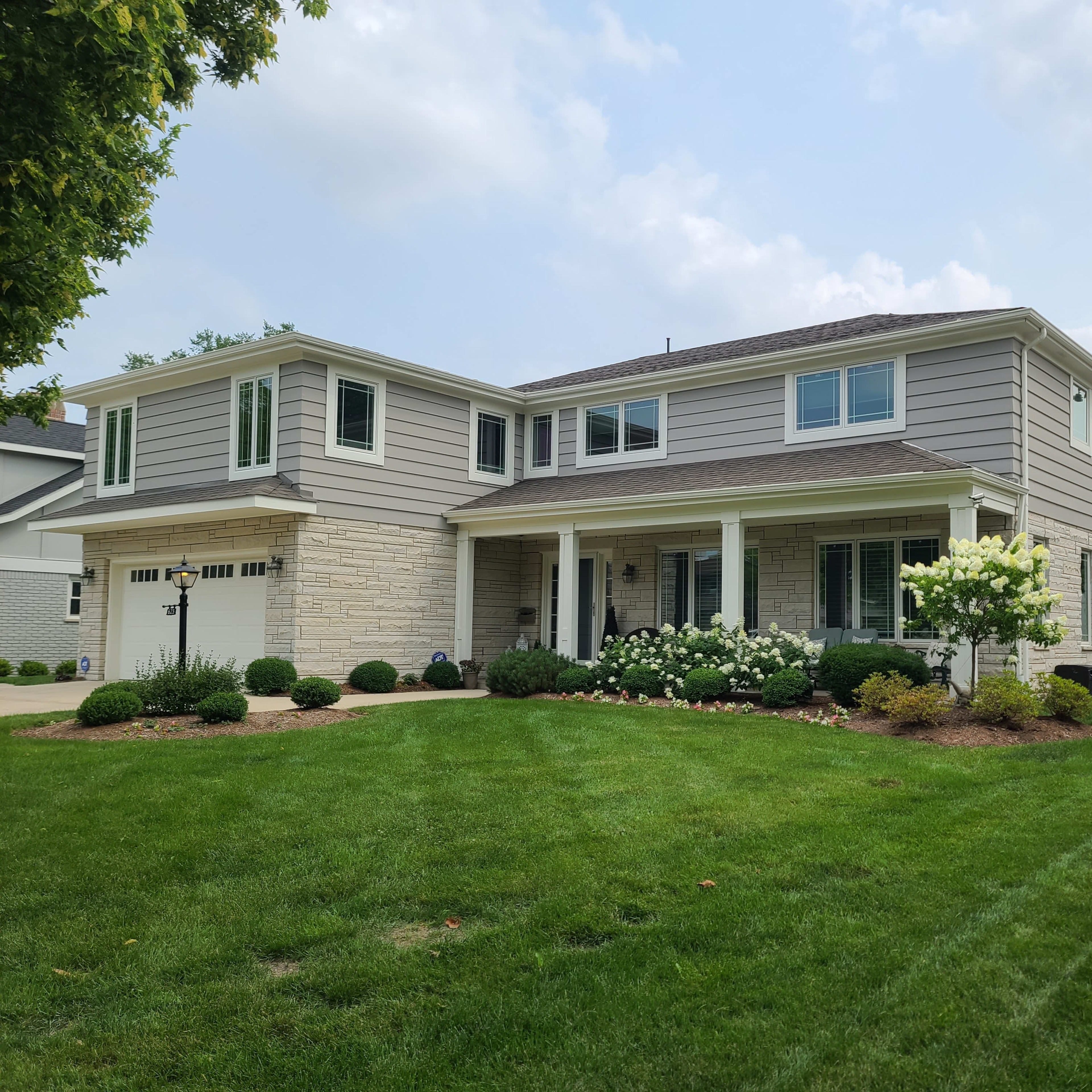 A two-story gray house with cream-colored stone accents and a well-maintained lawn featuring shrubs and flowers.