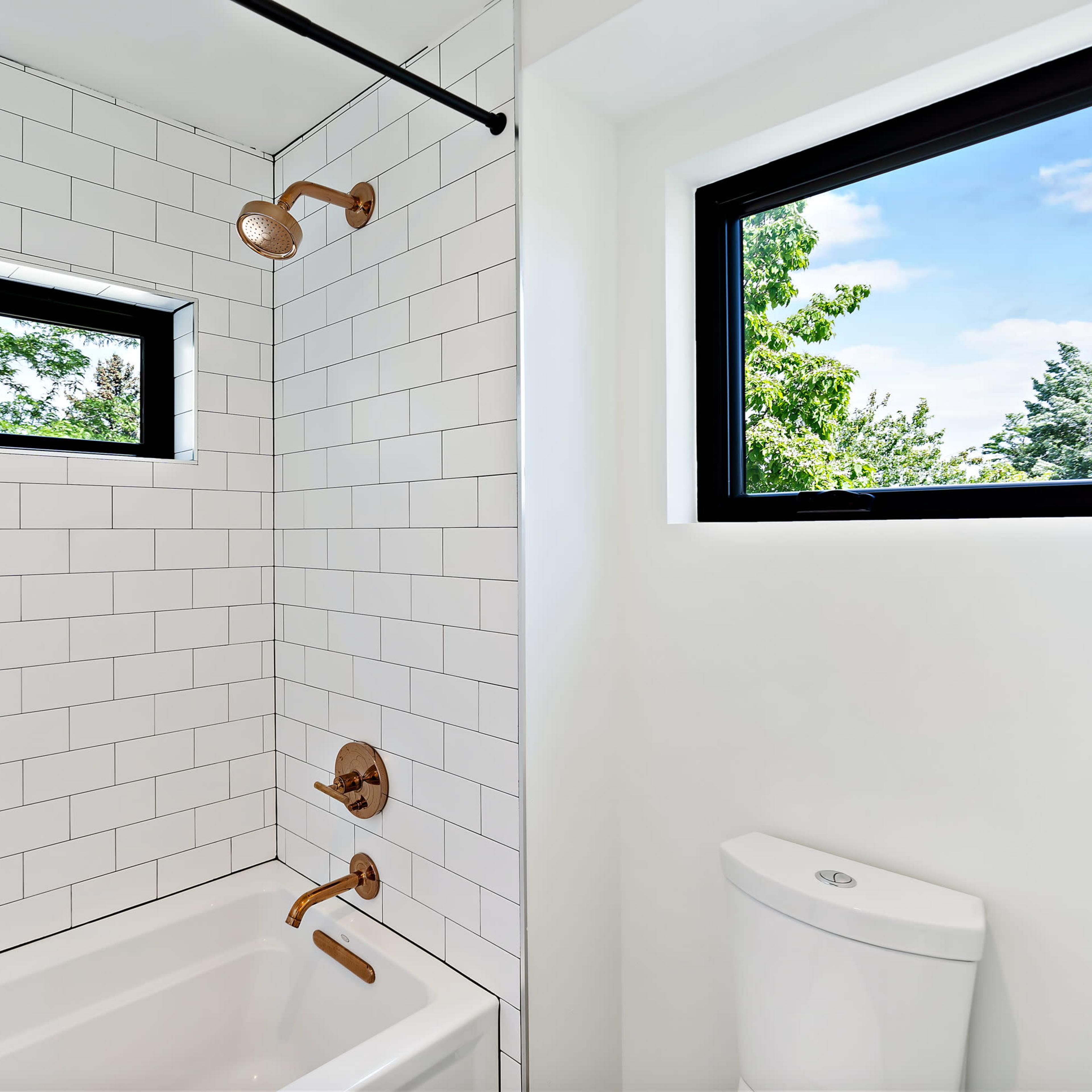 A bathroom with a white tiled shower area, a bathtub, and a modern toilet next to two windows that frame a view of greenery.