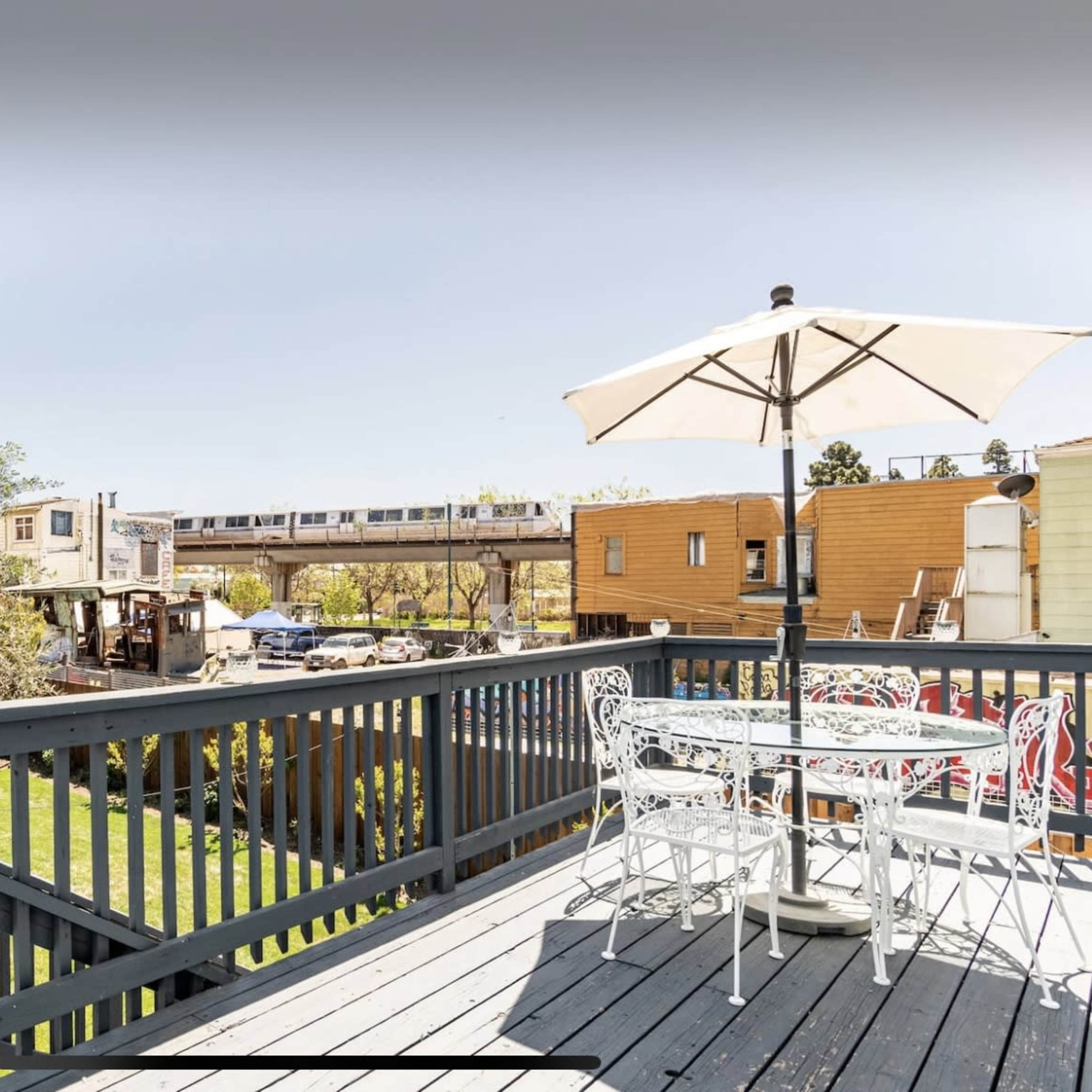 A wooden deck with a white table and chairs under an umbrella overlooks a neighborhood with buildings and a train passing nearby.