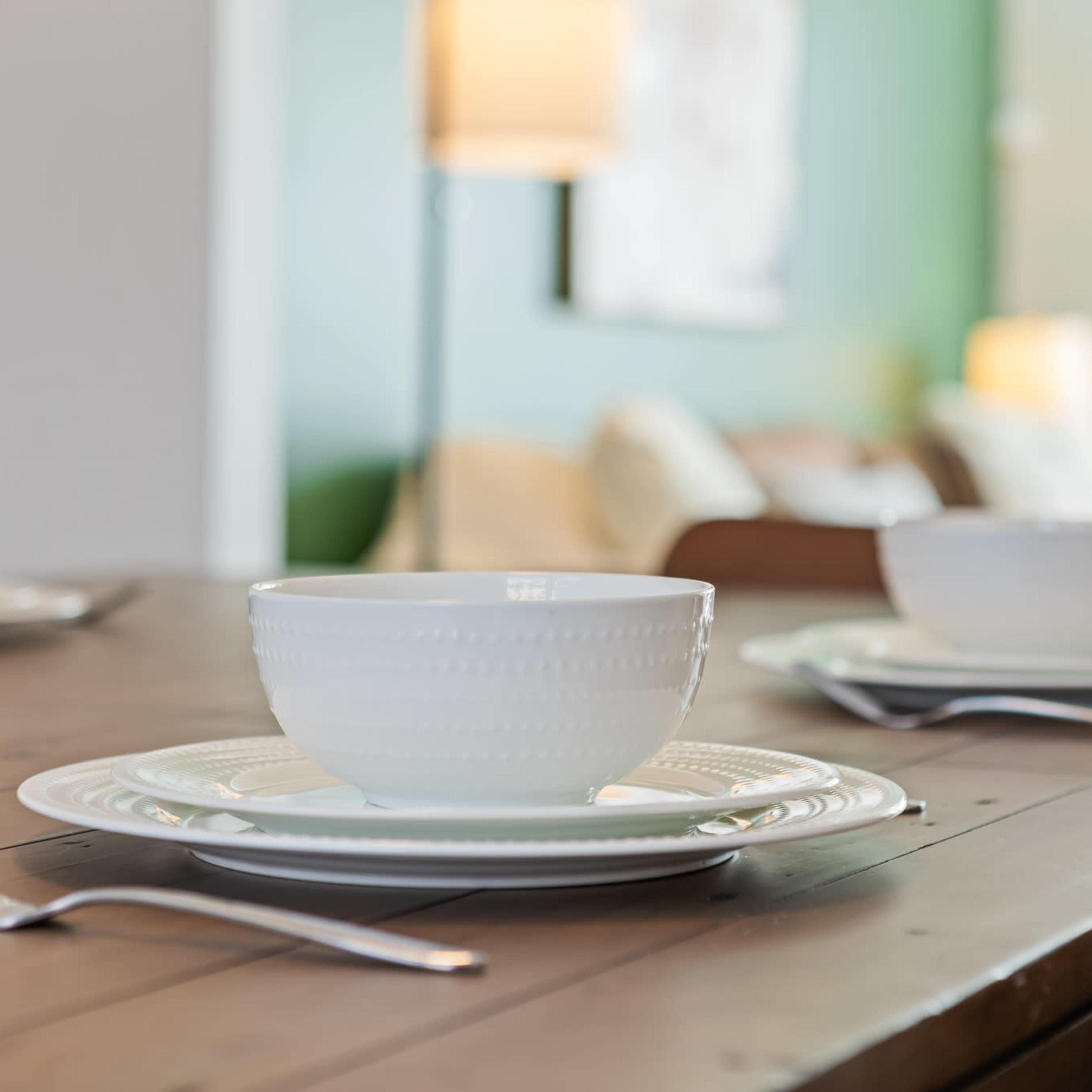 A wooden dining table is set with white plates and bowls, accompanied by stainless steel forks, in a well-lit room featuring a green wall and soft furnishings in the background.