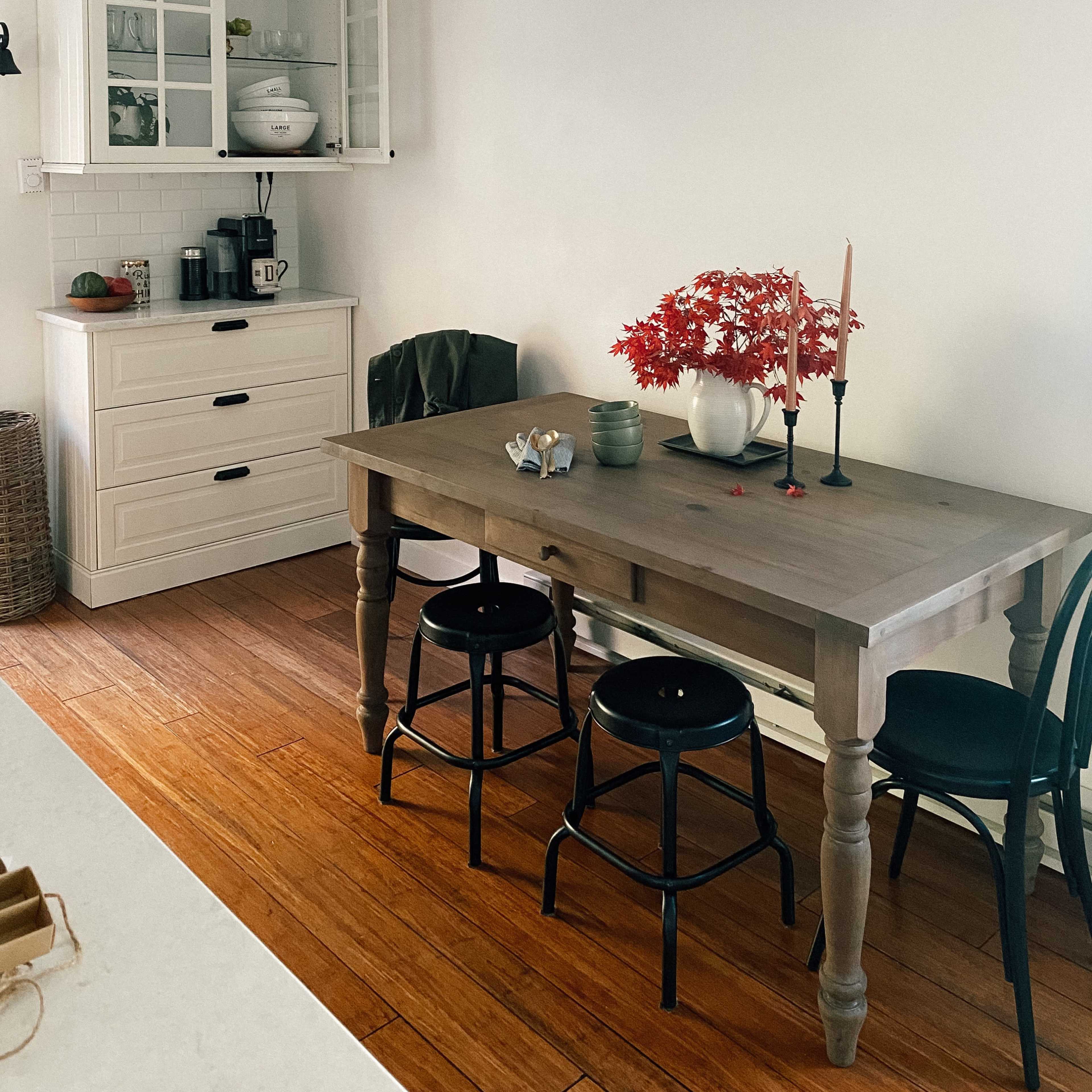 A wooden table with three black stools is set in a kitchen with a wall cabinet and a variety of decor items, including a vase with red foliage and candles.