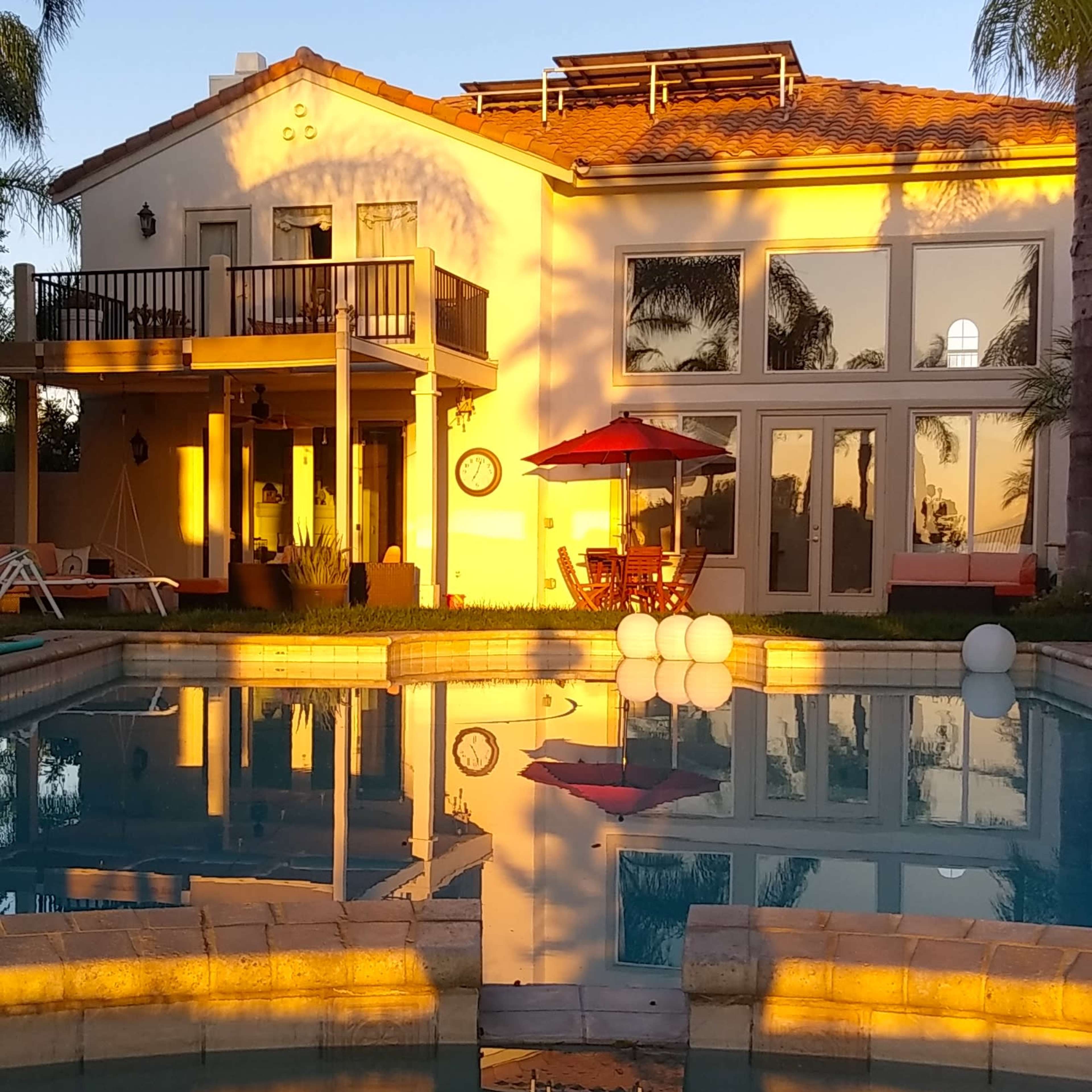 A large house with multiple balconies and a swimming pool reflects the evening light, surrounded by palm trees and lounge chairs.