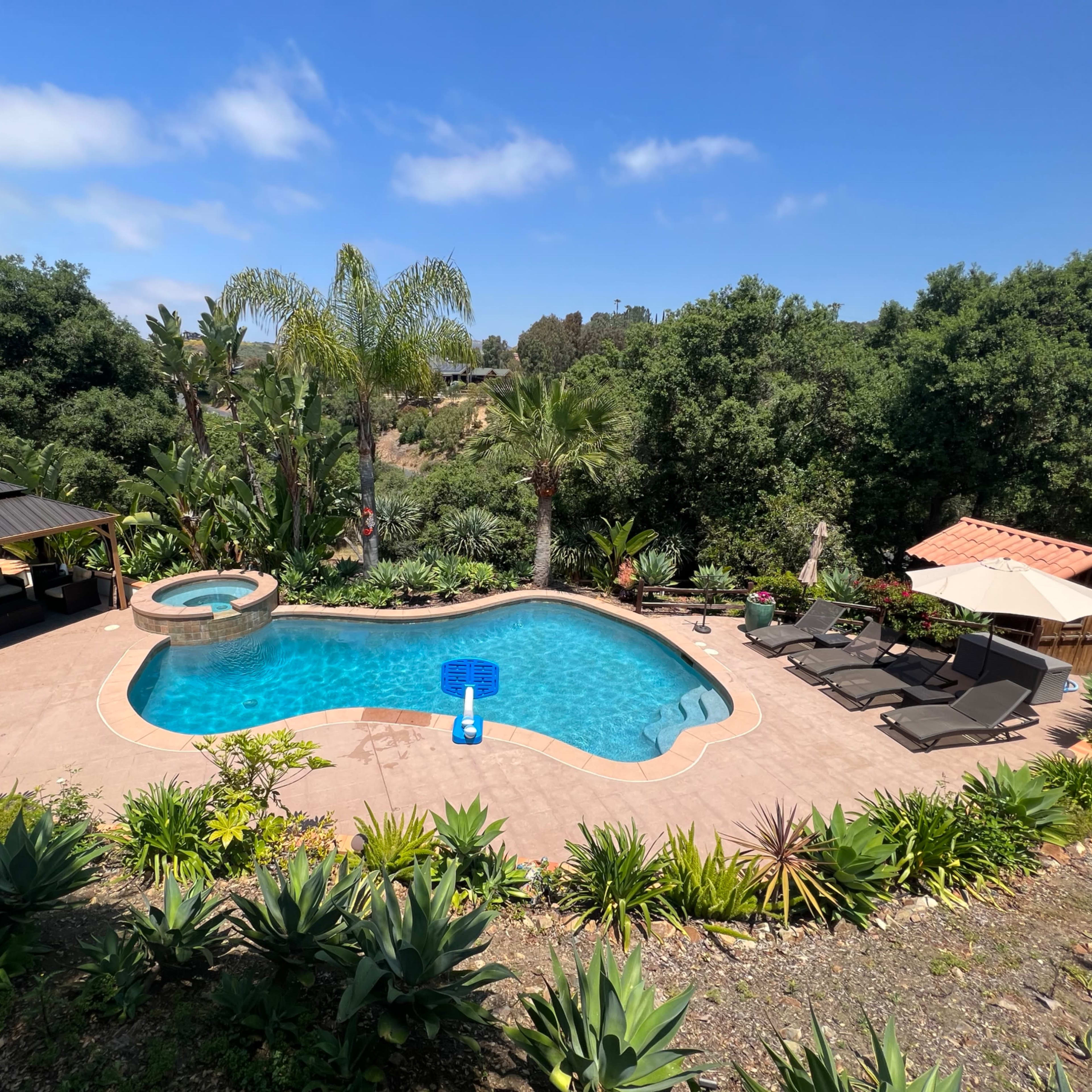 A backyard area with a swimming pool, hot tub, lounge chairs, and a gazebo surrounded by lush greenery.