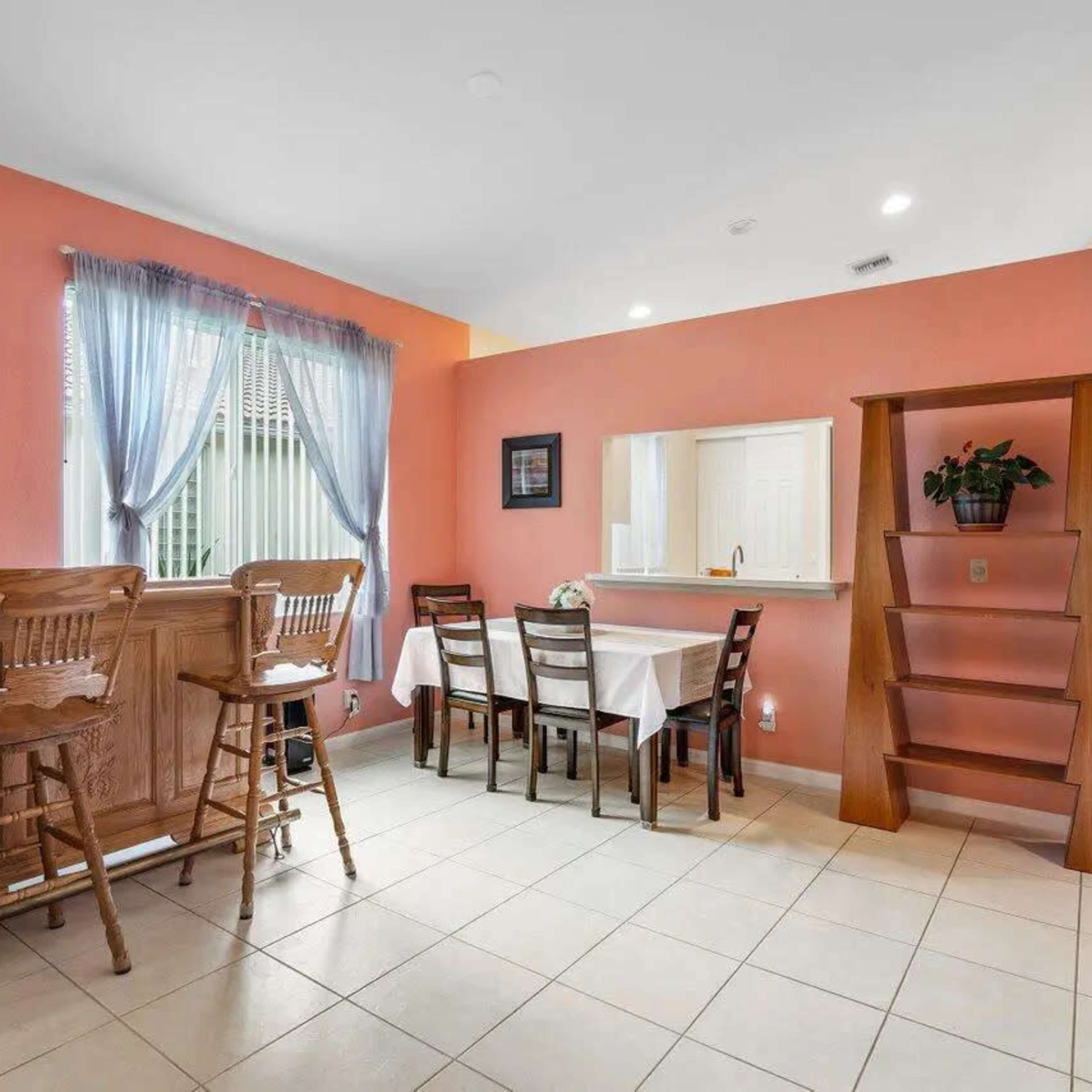 A dining area features a wooden bar with two stools, a table set for four, and a built-in shelf against a wall.