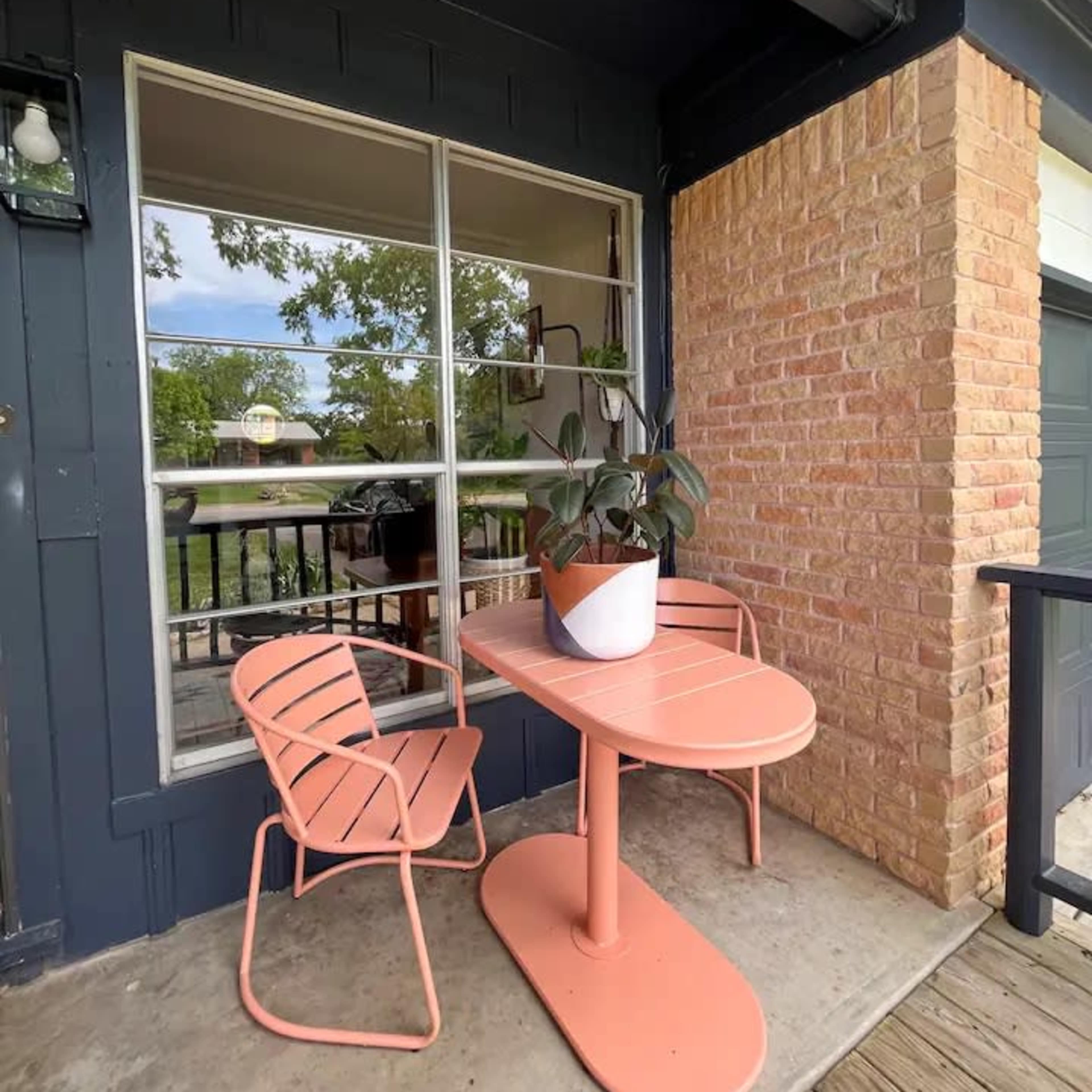 A small patio area features a pink table and two matching chairs, with a potted plant on the table and a large window in the background.