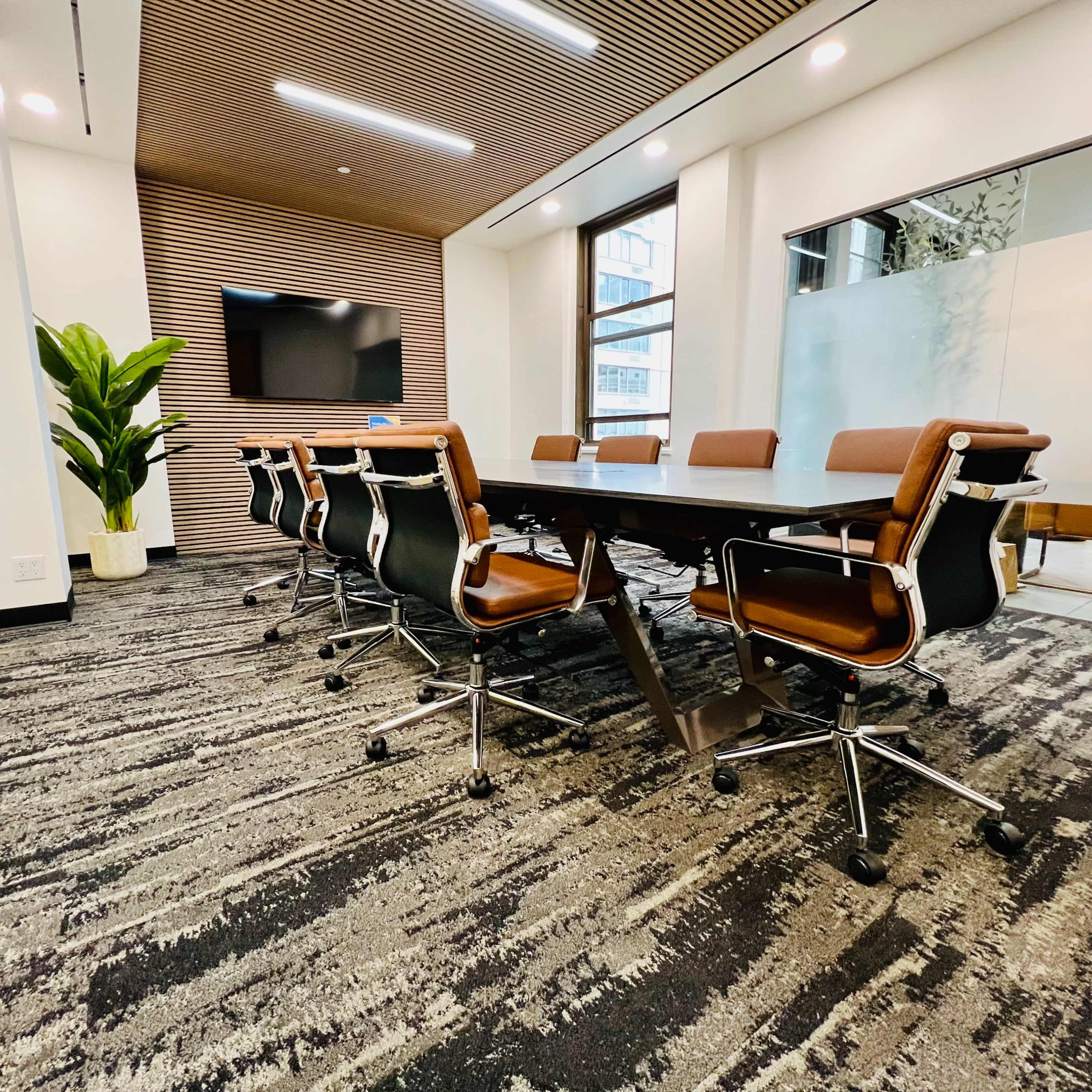 A modern conference room features a long table with rolling chairs, a large screen on the wall, and a plant in the corner.