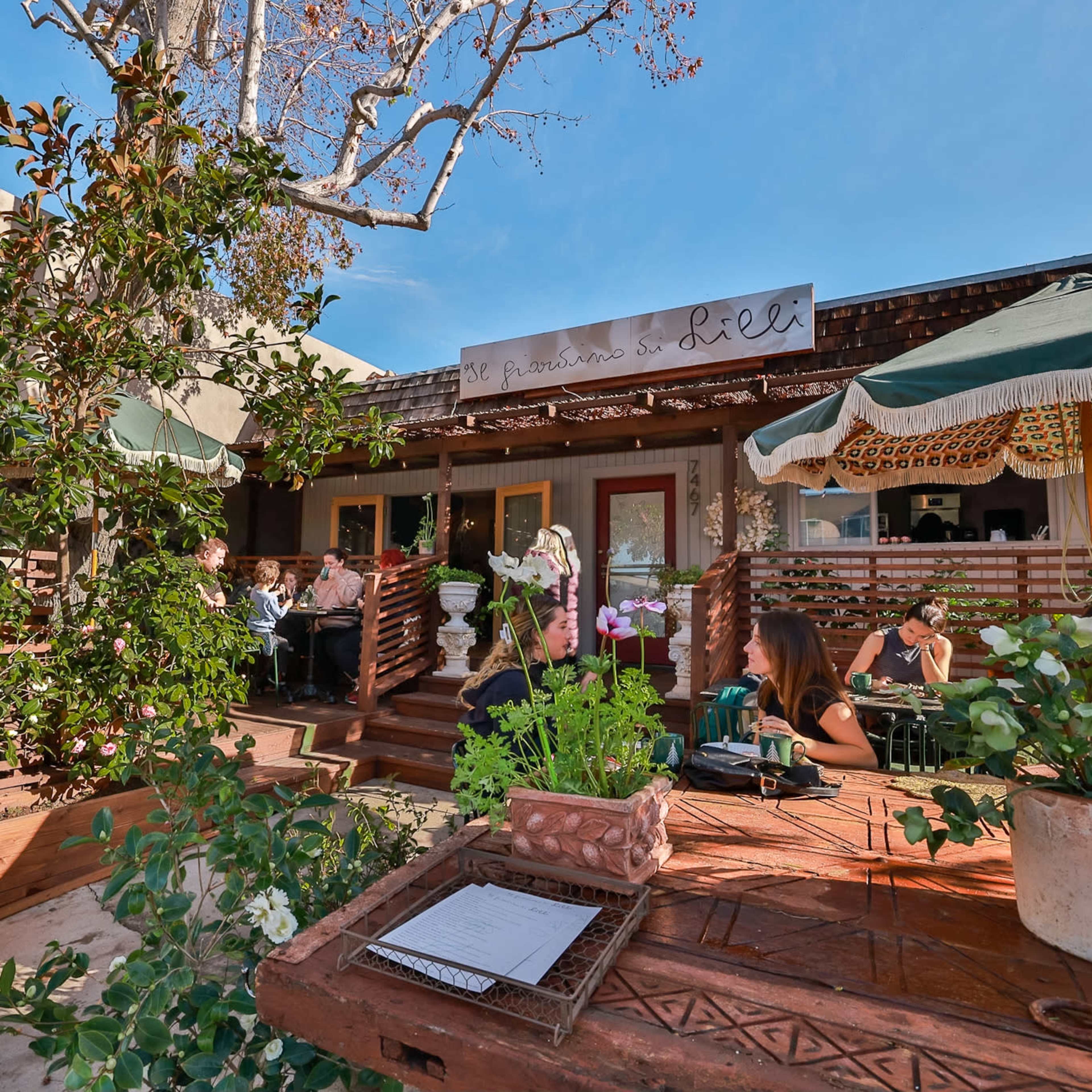 A restaurant patio features outdoor seating surrounded by greenery and canopies, with diners enjoying their meals under a clear blue sky.