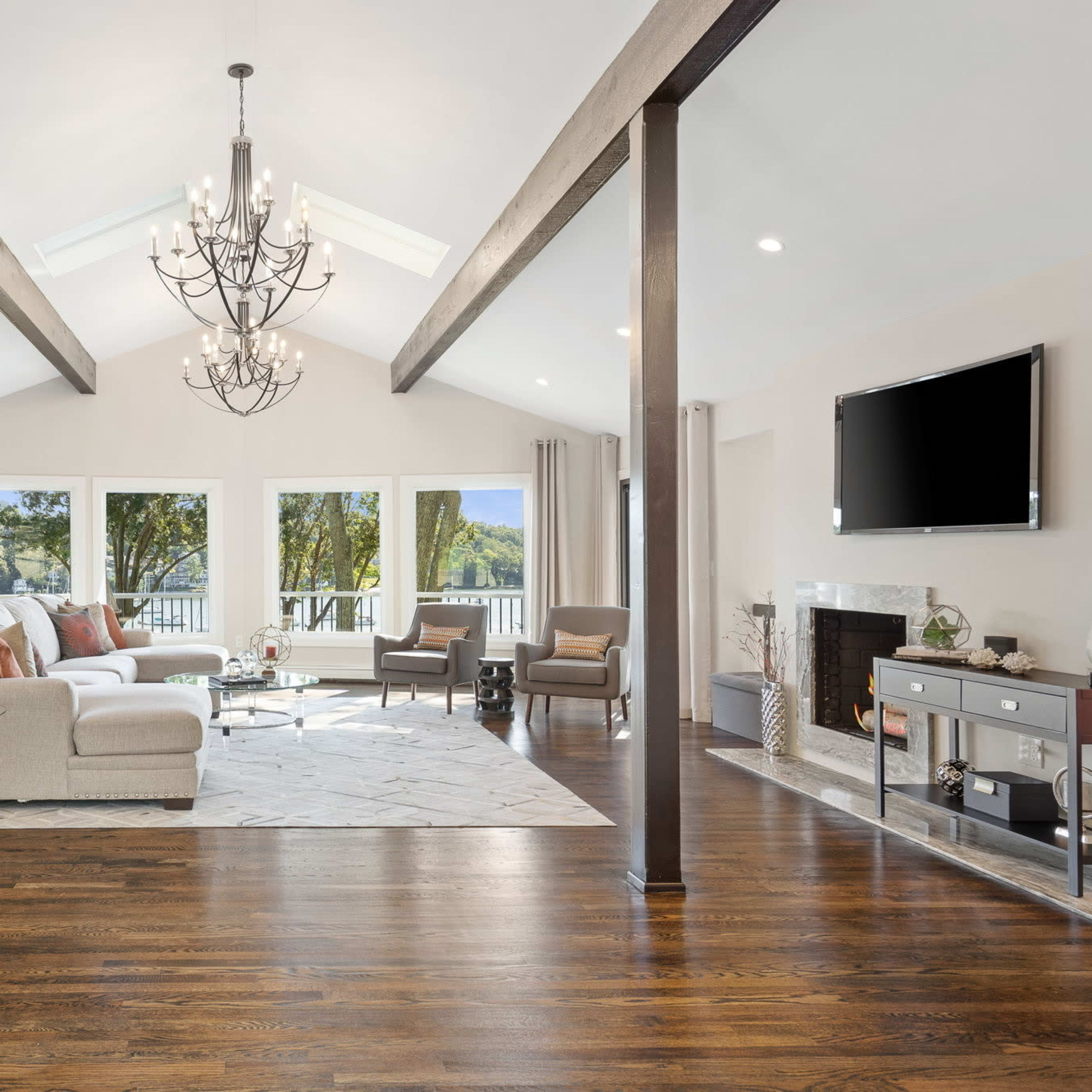 A spacious living room with large windows, wooden flooring, and modern furnishings, featuring a chandelier and a flat-screen TV.