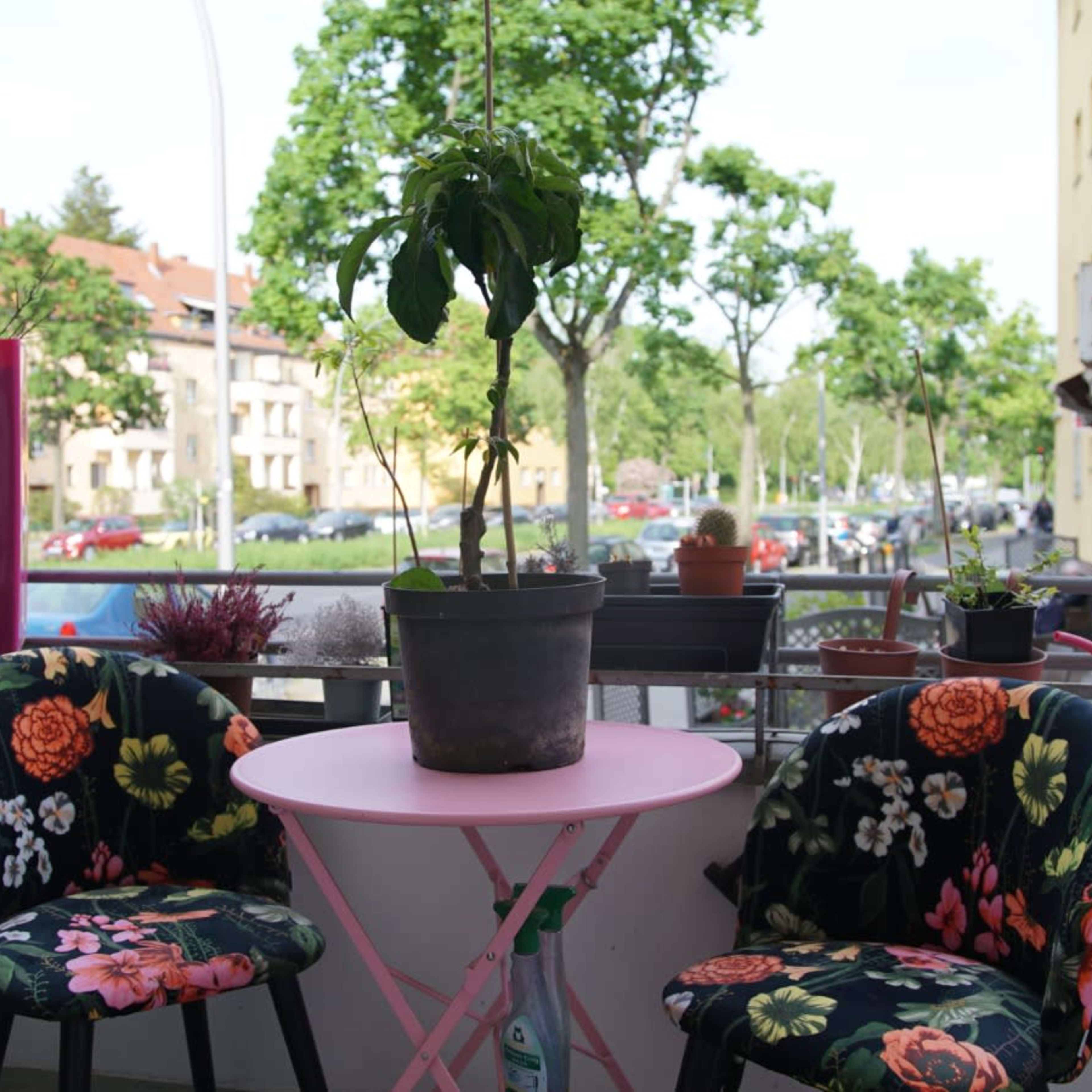 A small balcony features a round pink table, two floral-patterned chairs, and a potted plant, with a view of trees and buildings in the background.