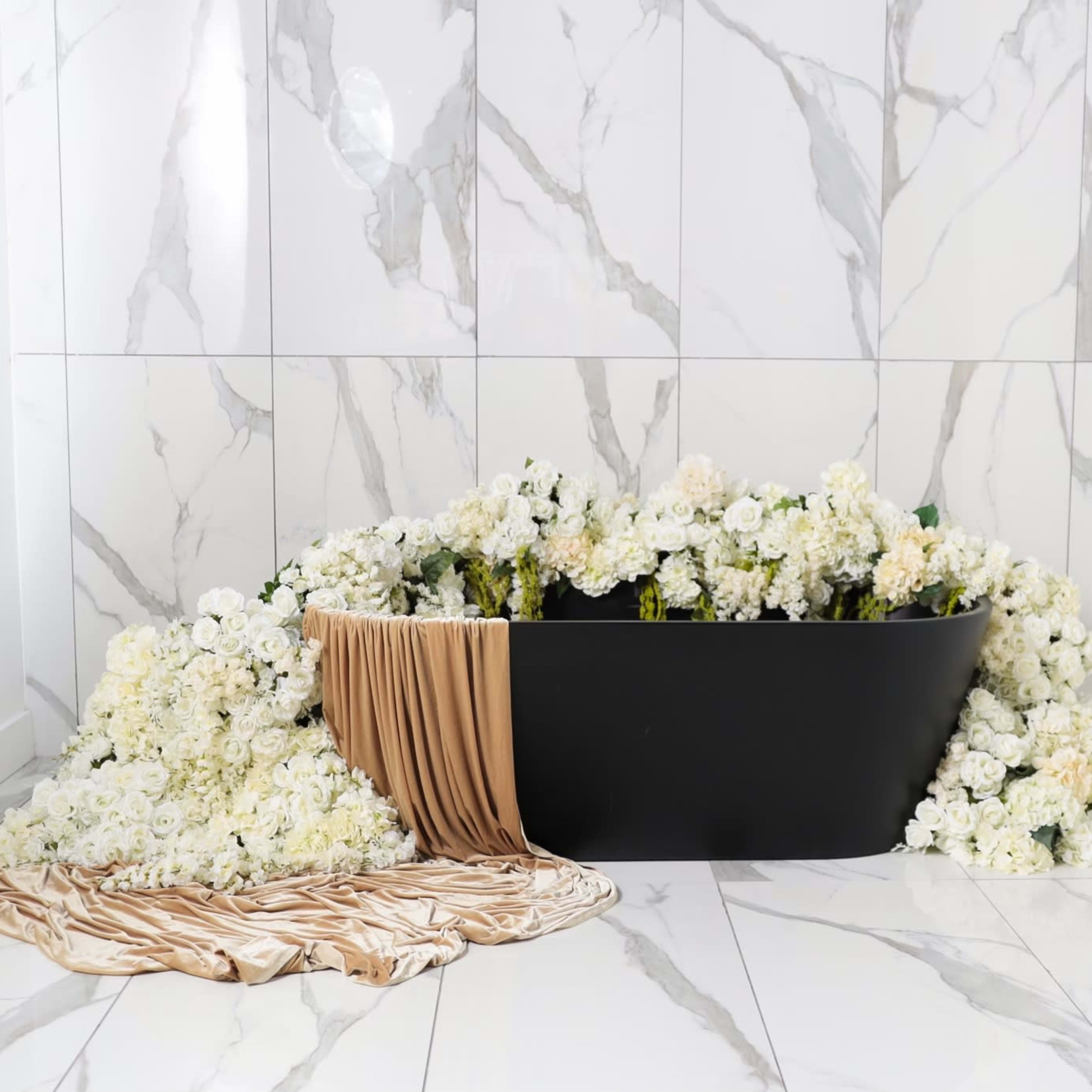 A black bathtub is surrounded by an array of white flowers against a marble wall.