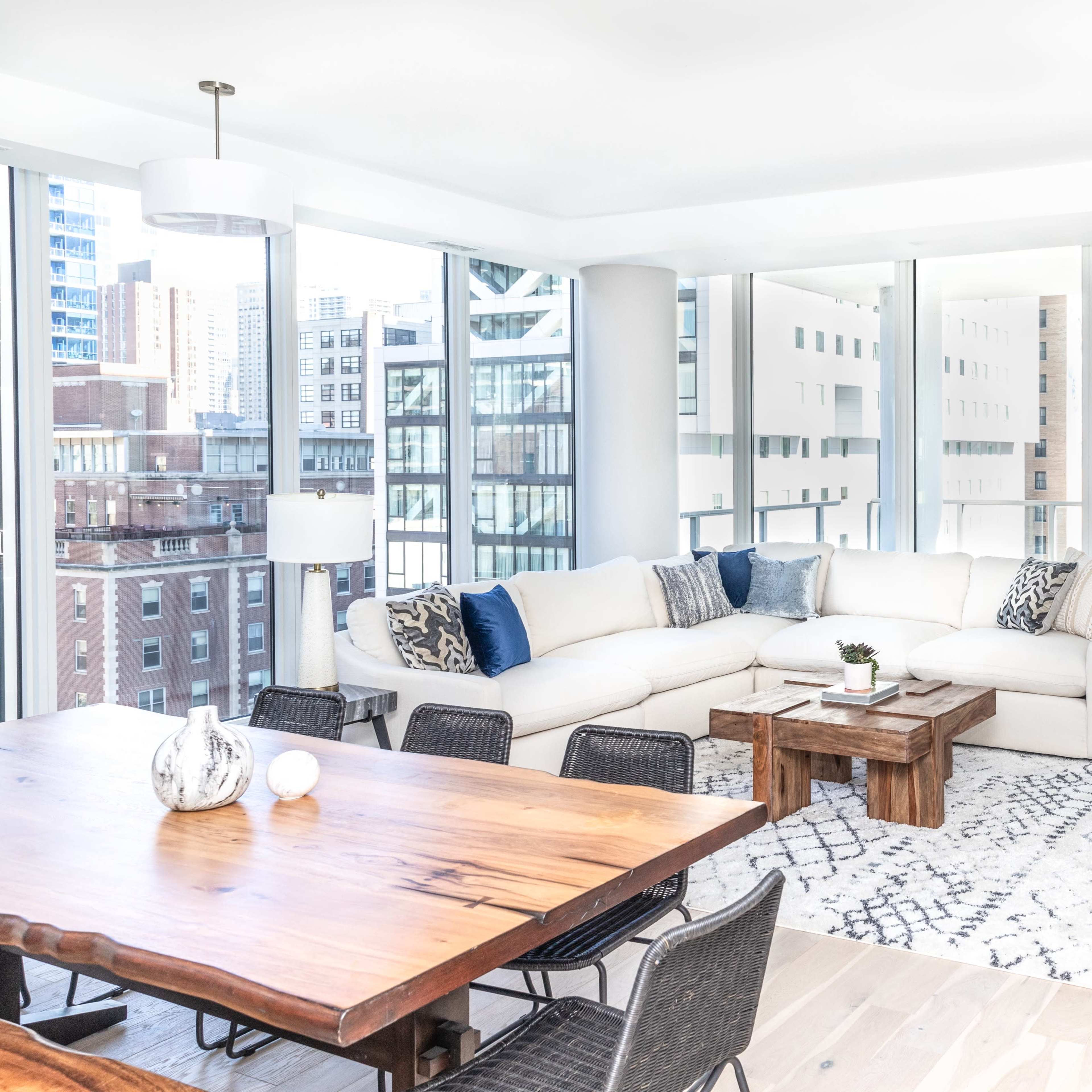 A modern living room features a large sectional sofa, a wooden coffee table, and a dining area with a wooden table, surrounded by large windows offering a city view.