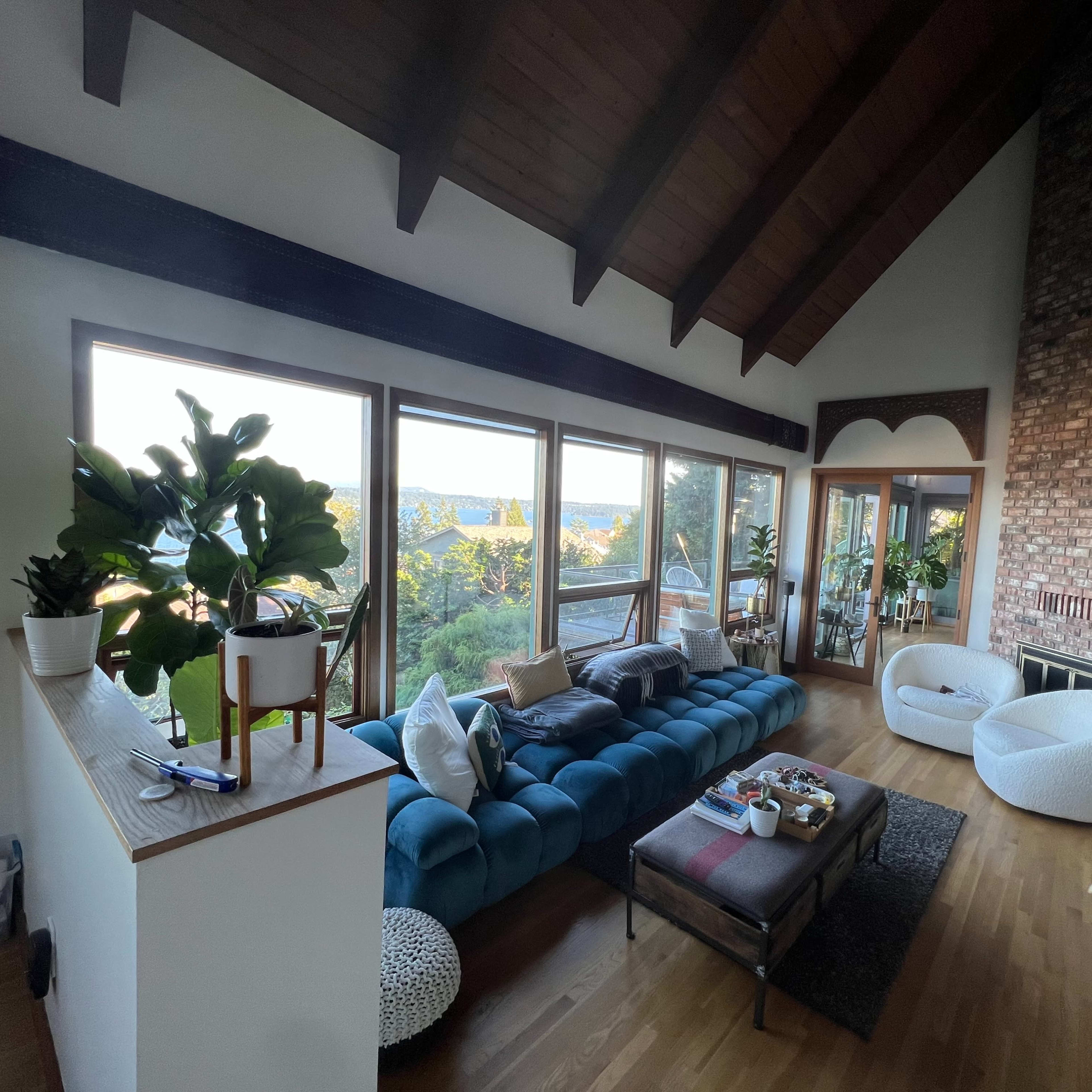 A spacious living room features a blue sofa, a coffee table, large windows with views of nature, and a high wooden ceiling.