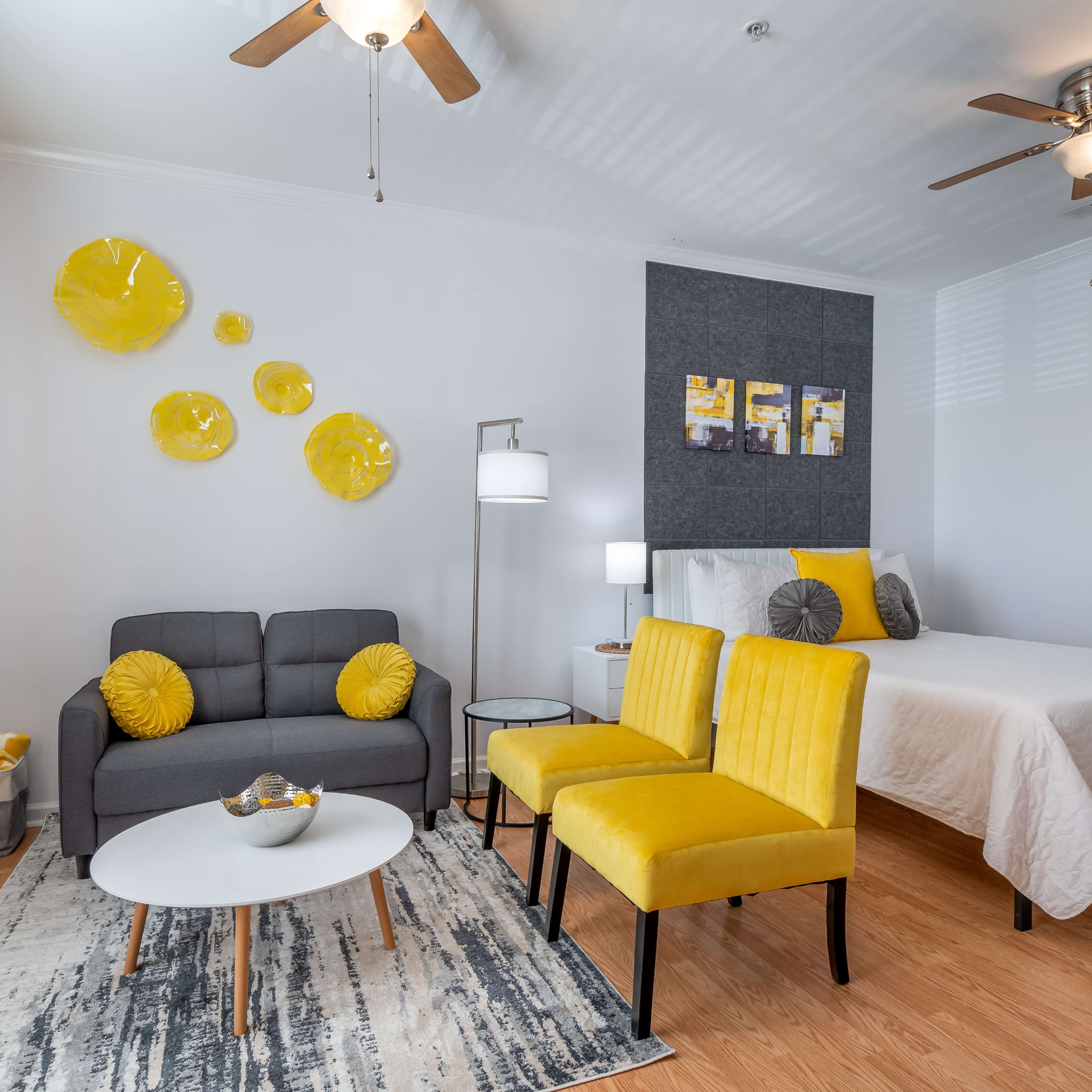 A modern, well-lit room featuring a bed, a gray sofa with yellow pillows, and two yellow chairs arranged around a round coffee table on a patterned rug.