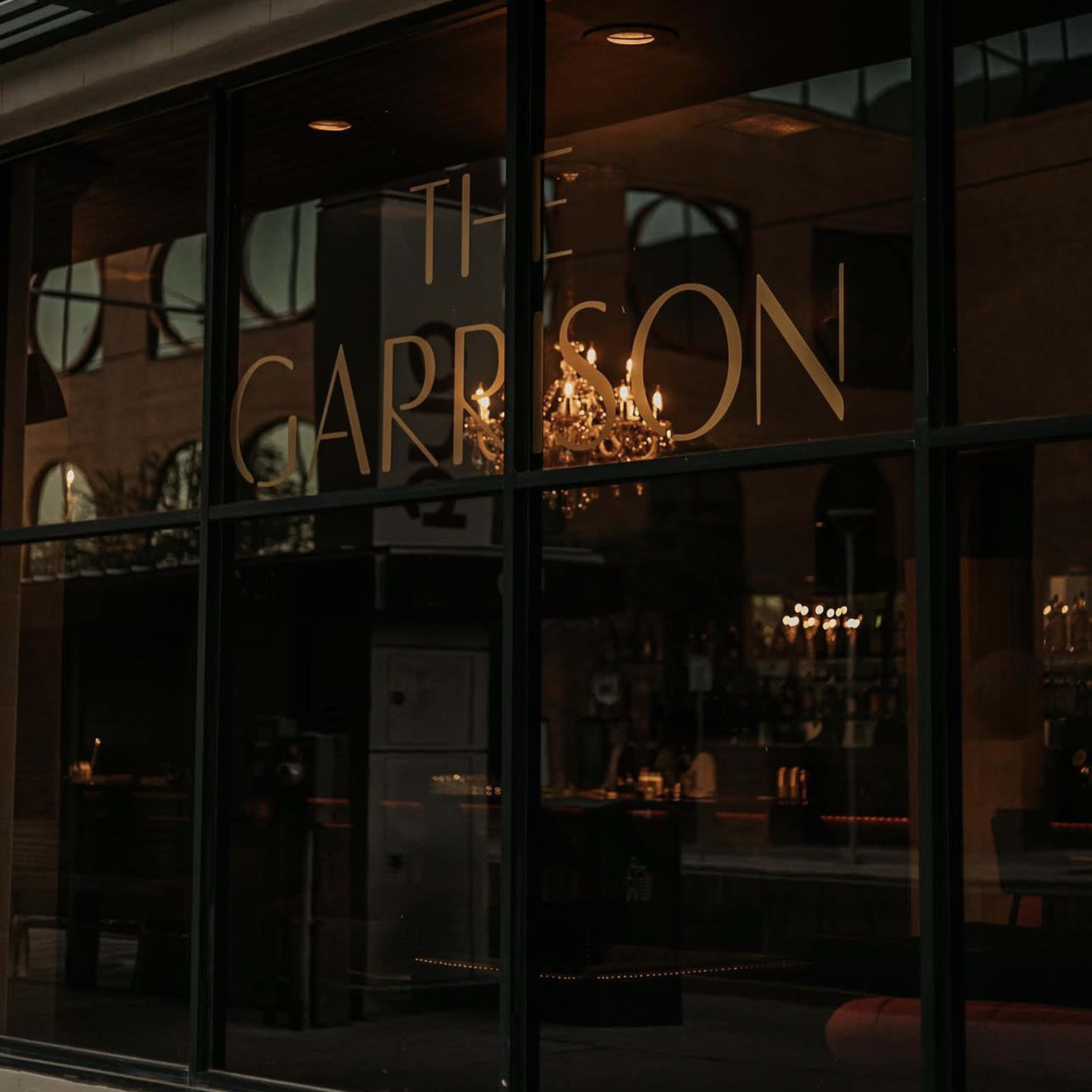 The entrance of a building with the sign "The Garrison" reflected in a large glass window, featuring a chandelier inside.