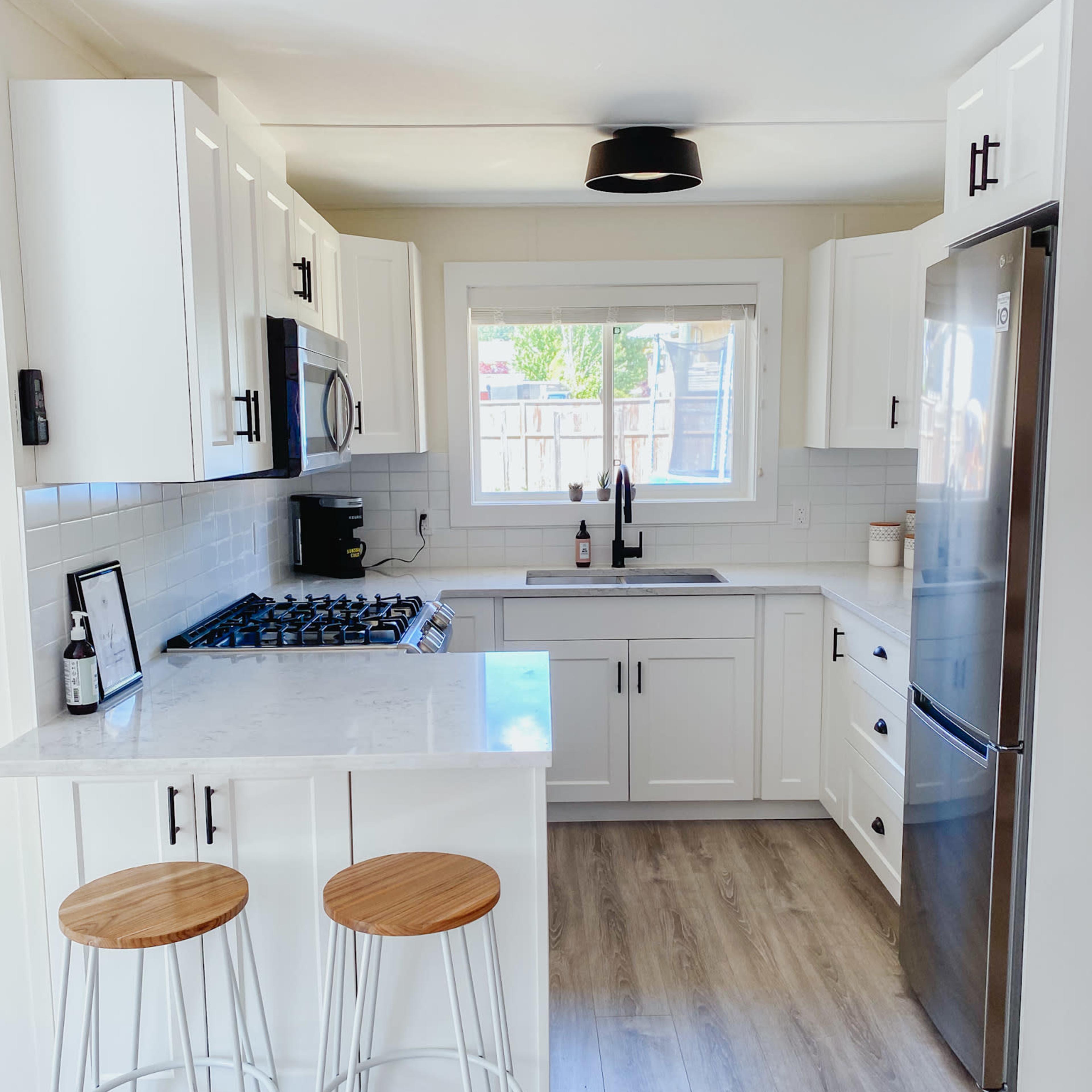 A modern kitchen featuring white cabinetry, a stainless steel refrigerator, a gas stove, and two bar stools at a light-colored countertop.