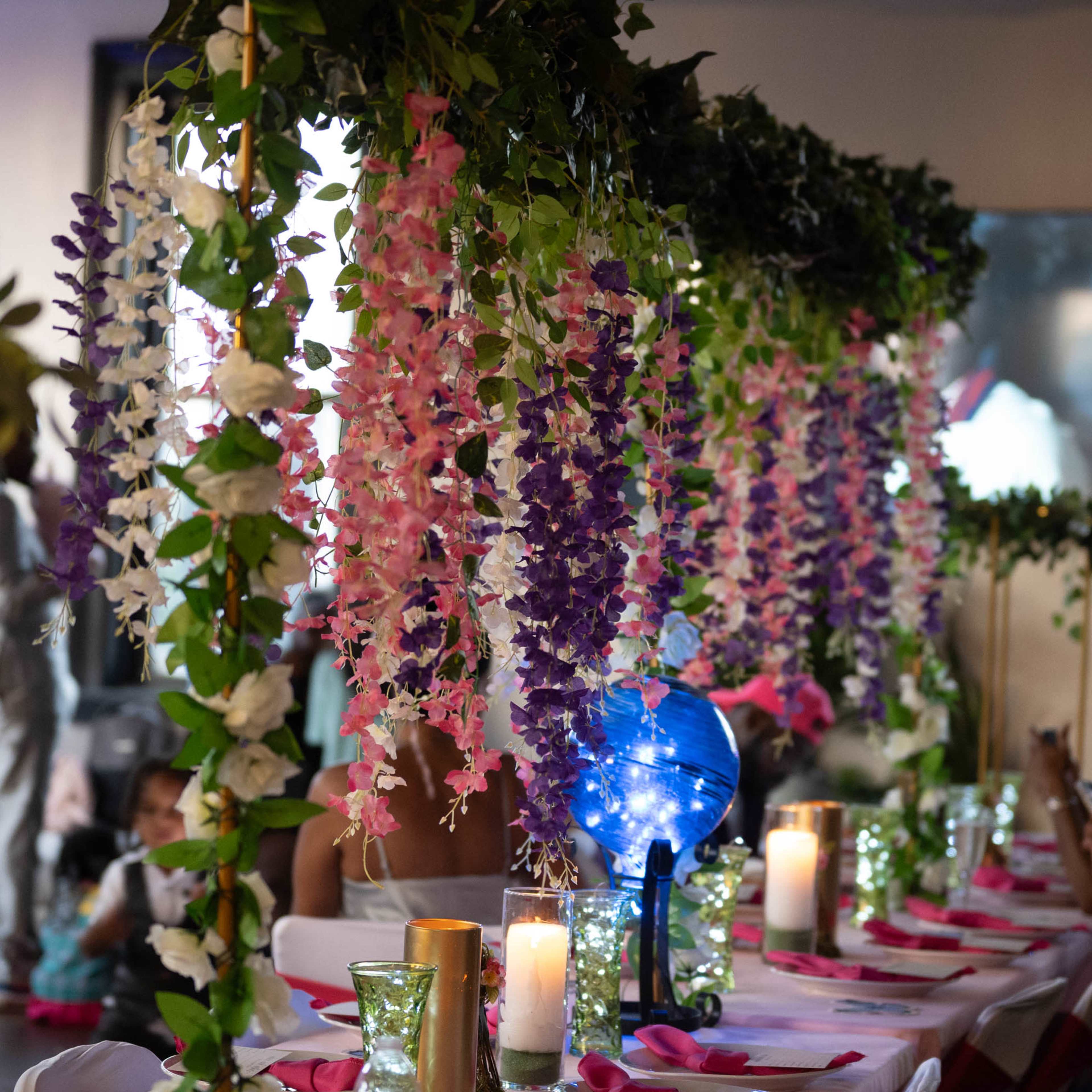 A decorated dining area features rows of flowers hanging from a canopy above tables set with candles and colorful tableware.