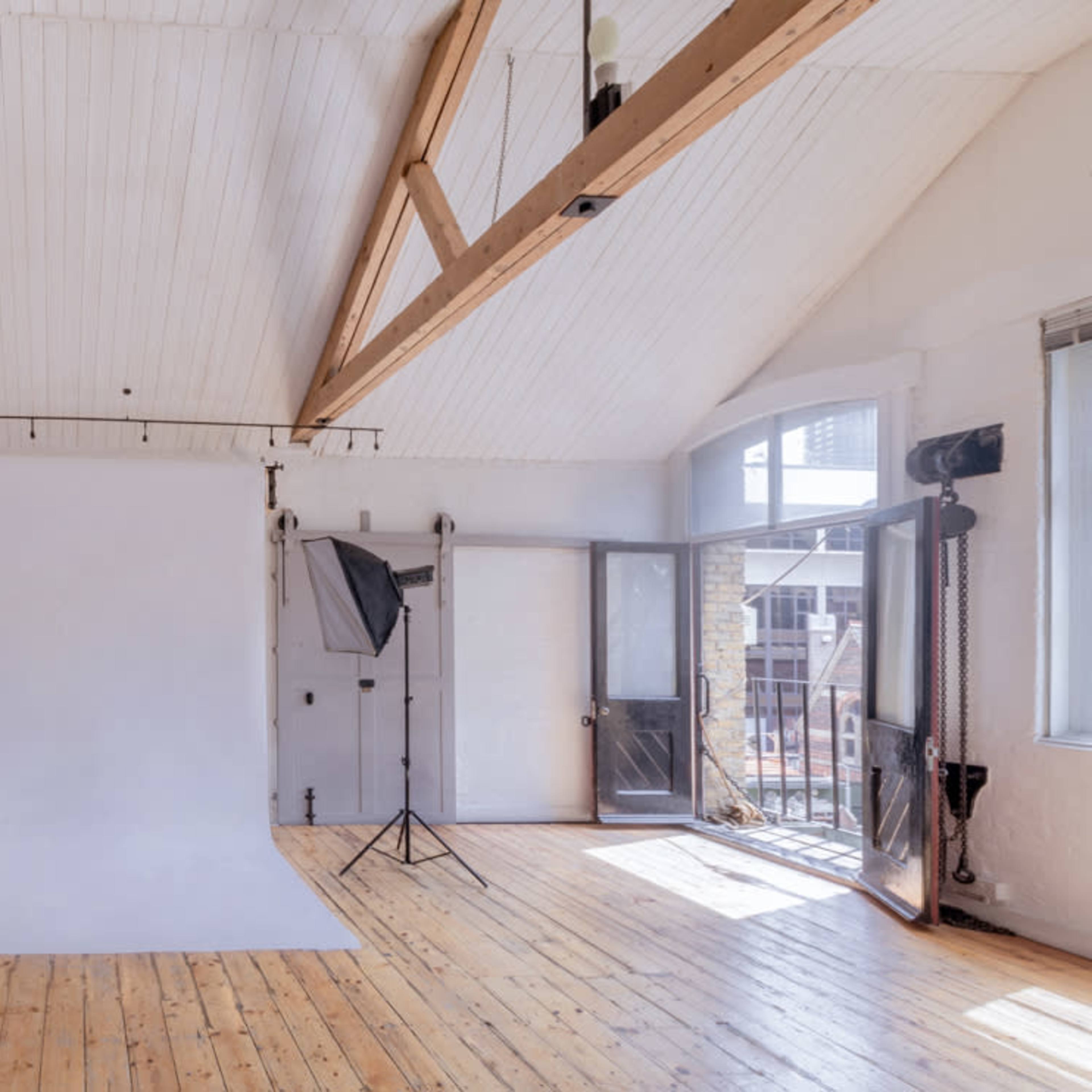 A photography studio with a white backdrop, wooden floors, and large windows, complemented by studio lighting equipment.