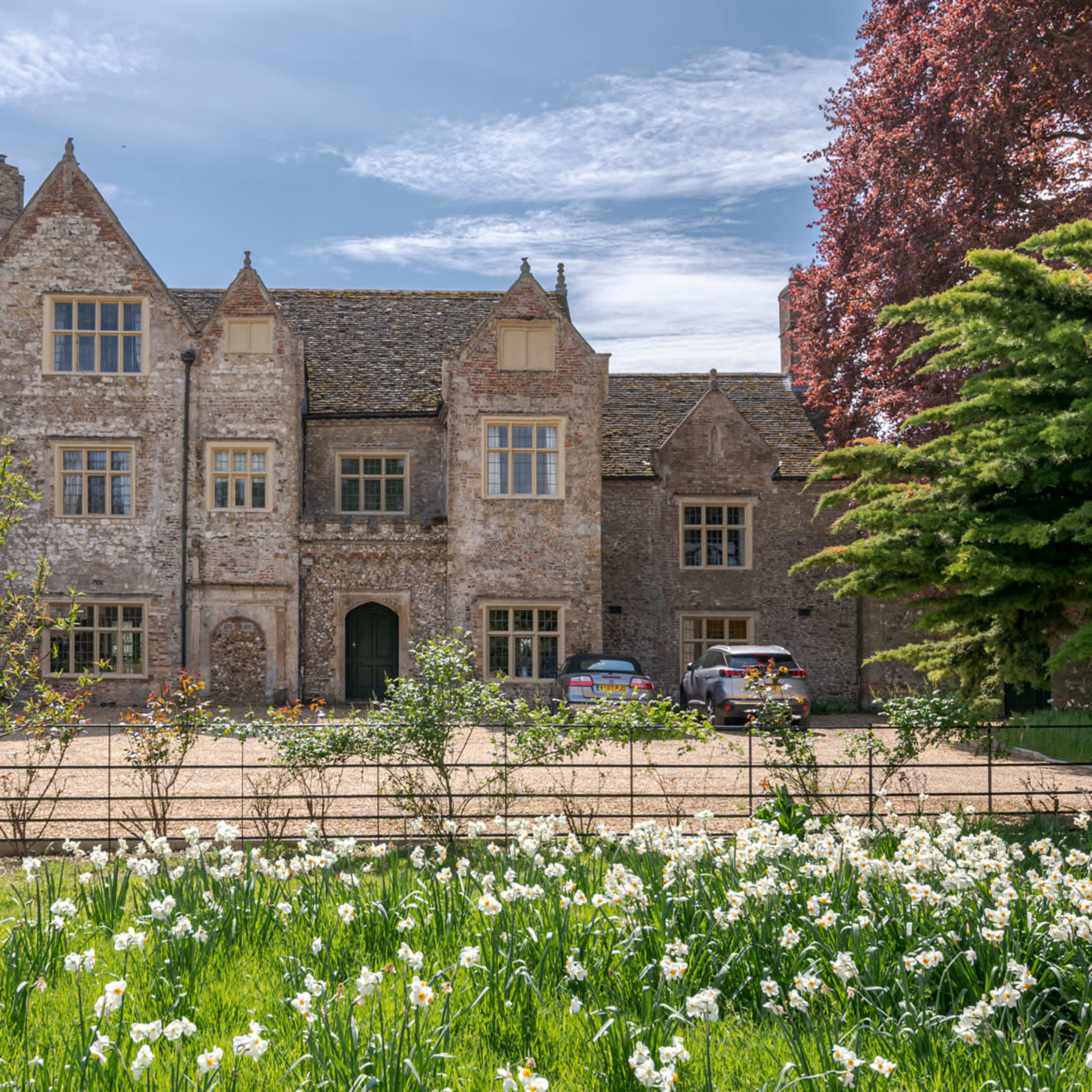 A stone manor house with a tower stands in front of a landscaped garden filled with daffodils.