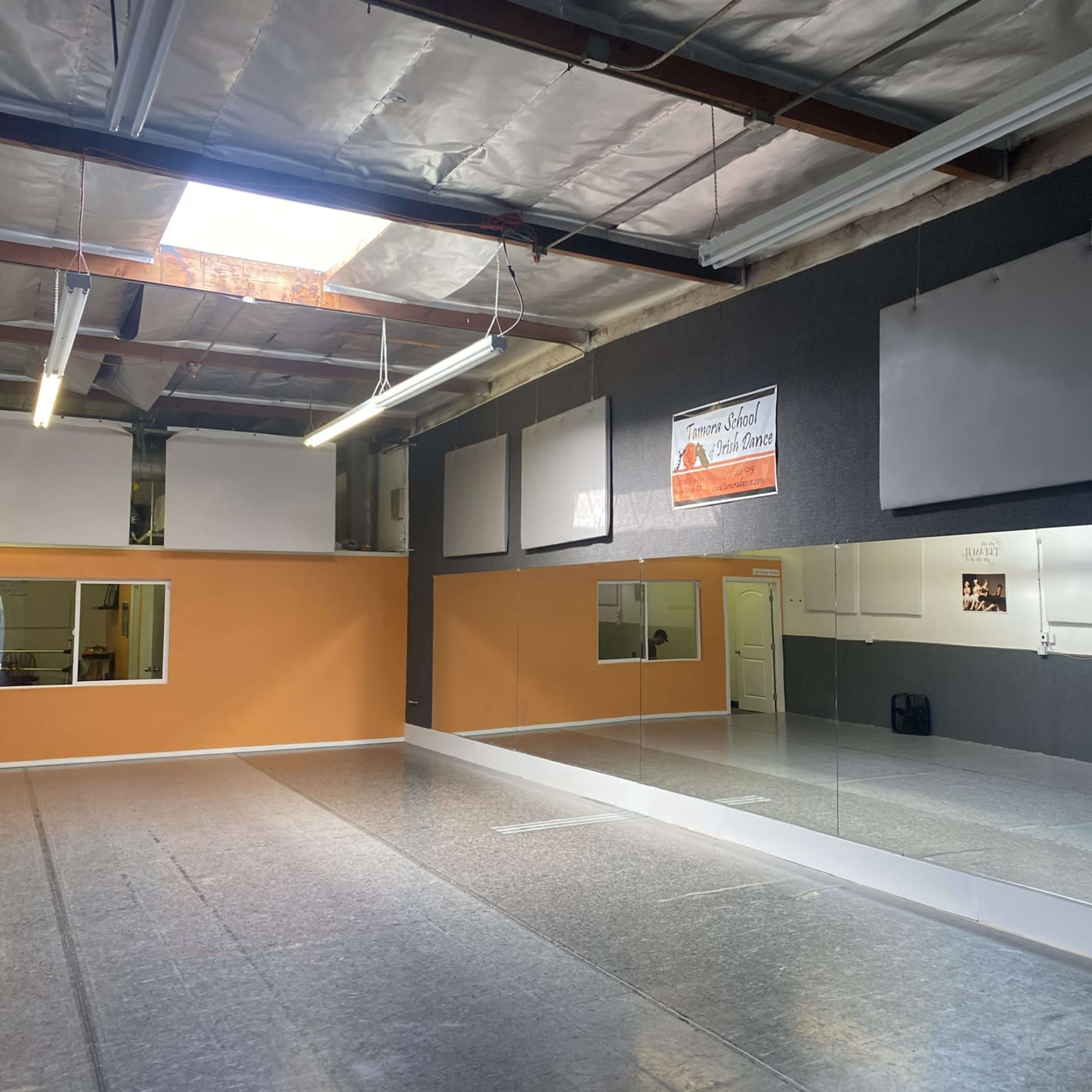 A dance studio with a large mirror on one wall, orange and gray walls, and overhead fluorescent lighting.