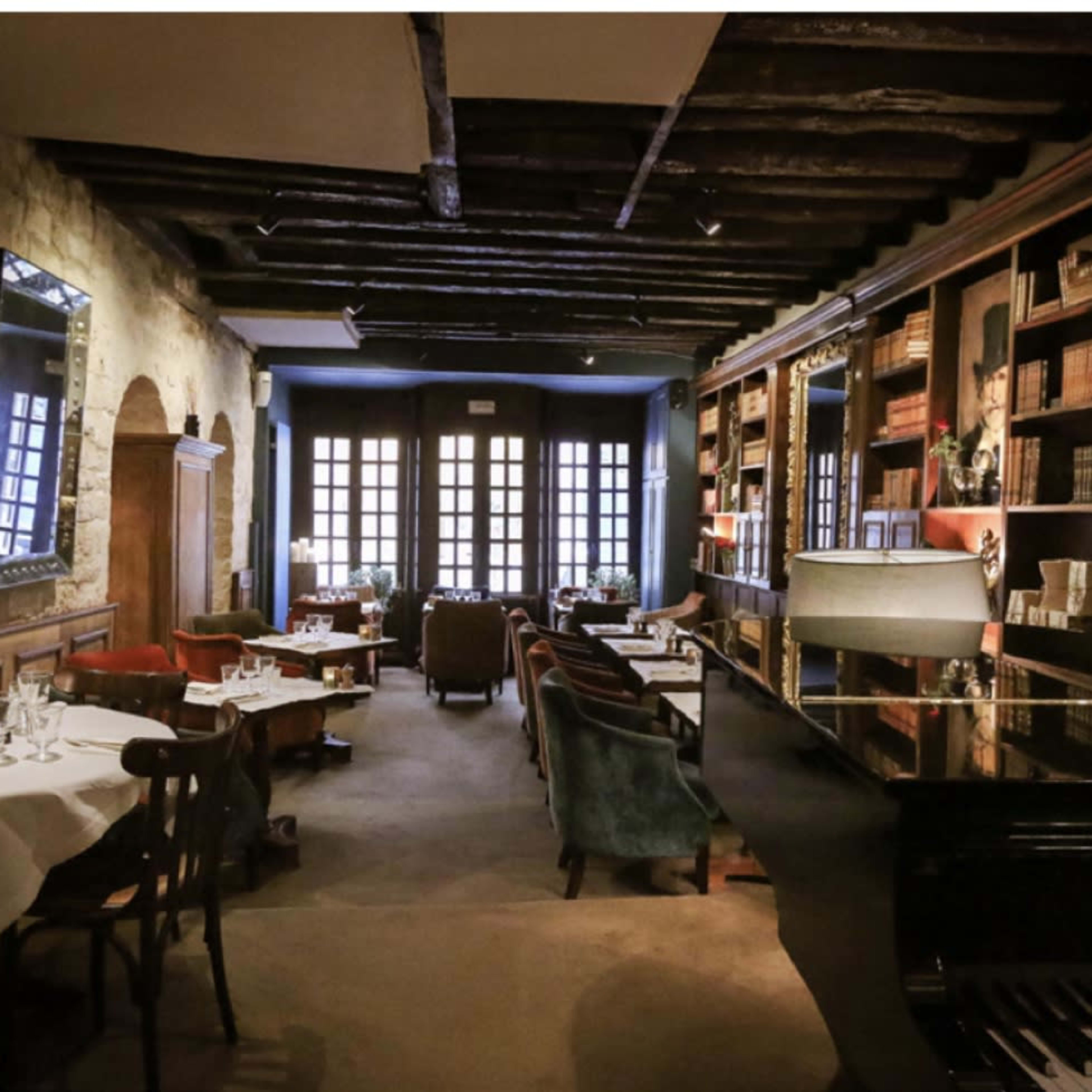 A dimly lit restaurant with wooden beams, tables set for dining, bookshelves lining the walls, and a piano in the corner.