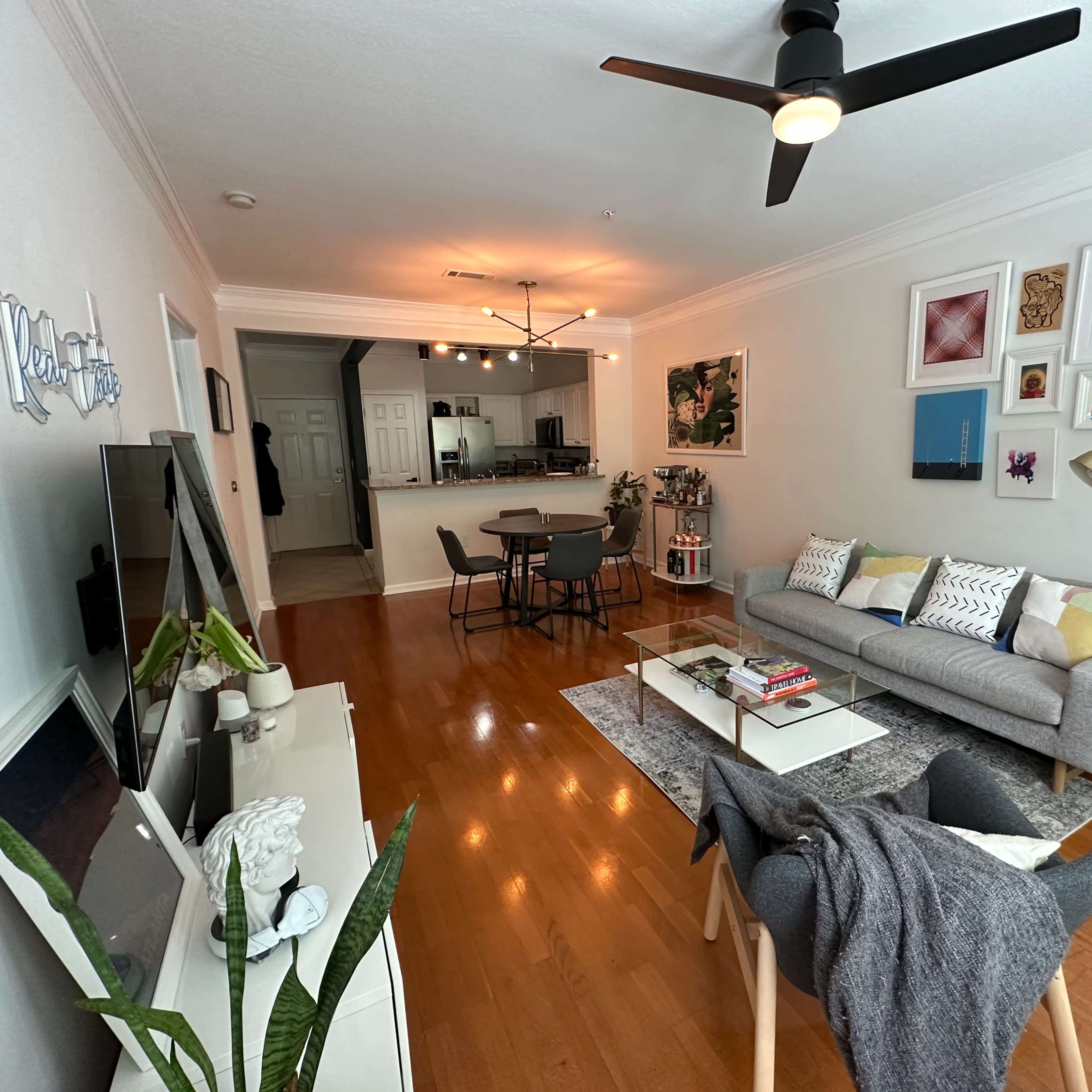 A living room features a grey sofa, a coffee table with books, a dining area in the background, and various artwork on the walls.