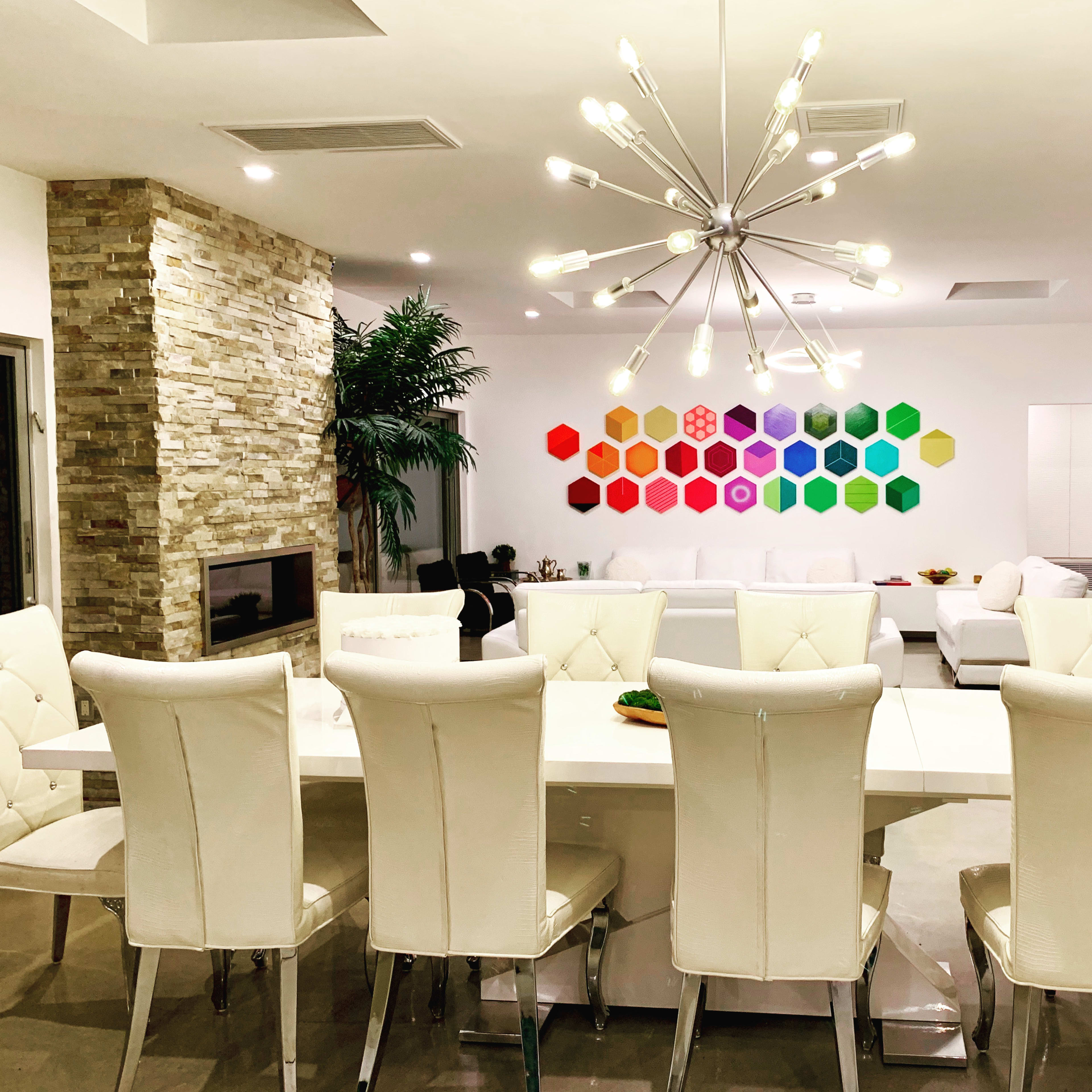 A modern living space featuring a long white dining table with eight white chairs, a colorful hexagon art installation on the wall, and a cozy seating area with a fireplace.