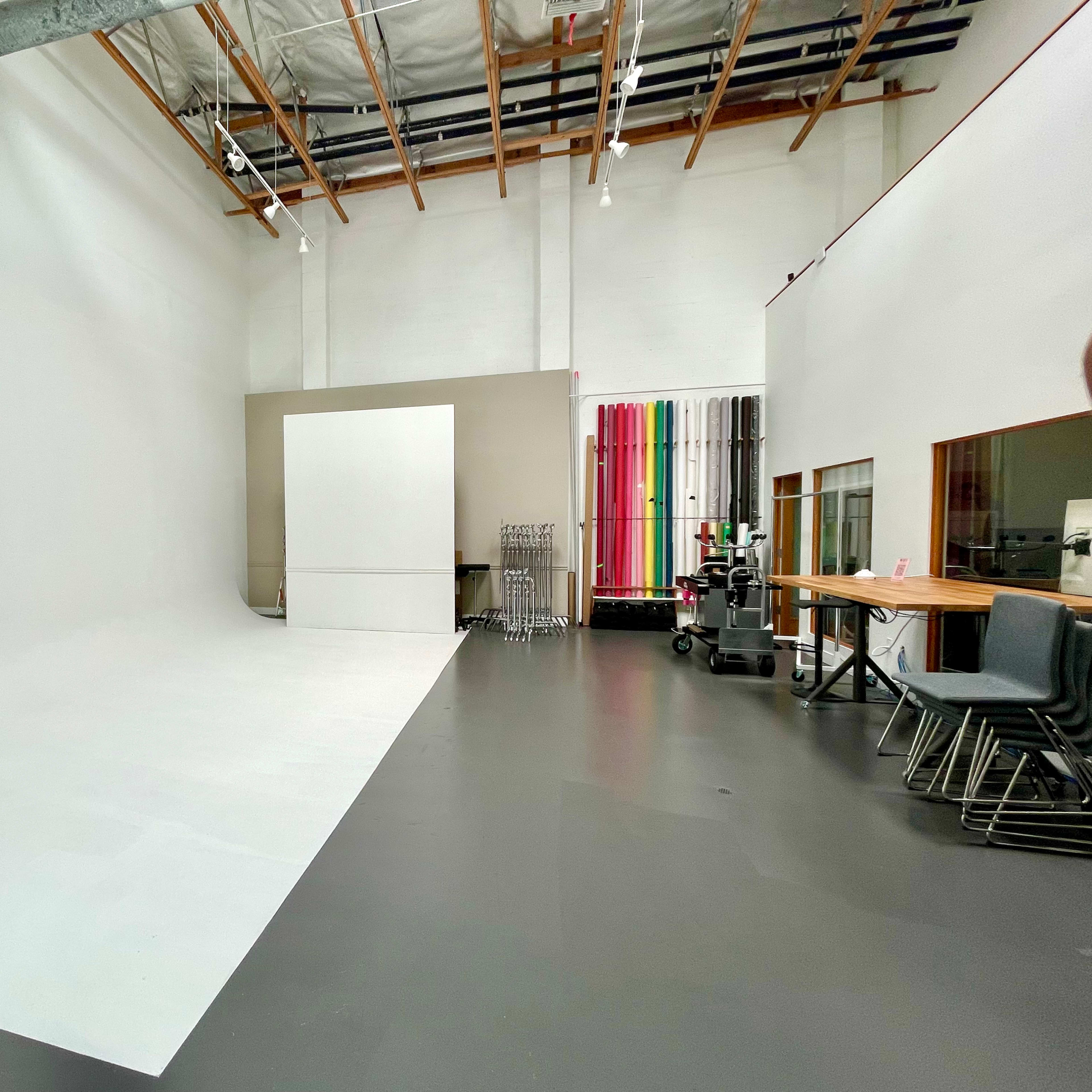 A spacious photography studio with a white backdrop, gray flooring, and colorful backdrops neatly arranged against the wall.