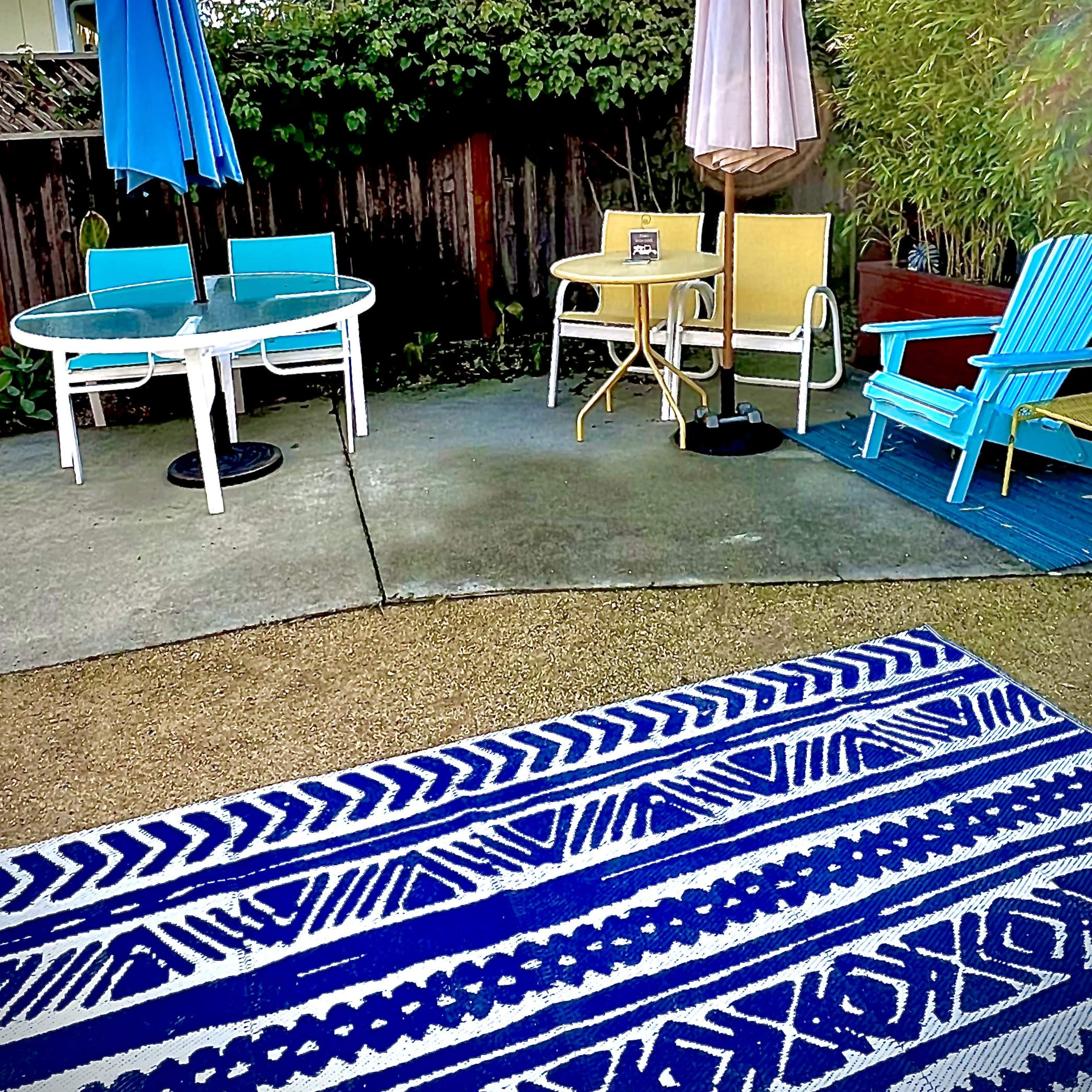 A backyard scene with various chairs and tables arranged on a concrete patio, surrounded by greenery and decorative lighting.