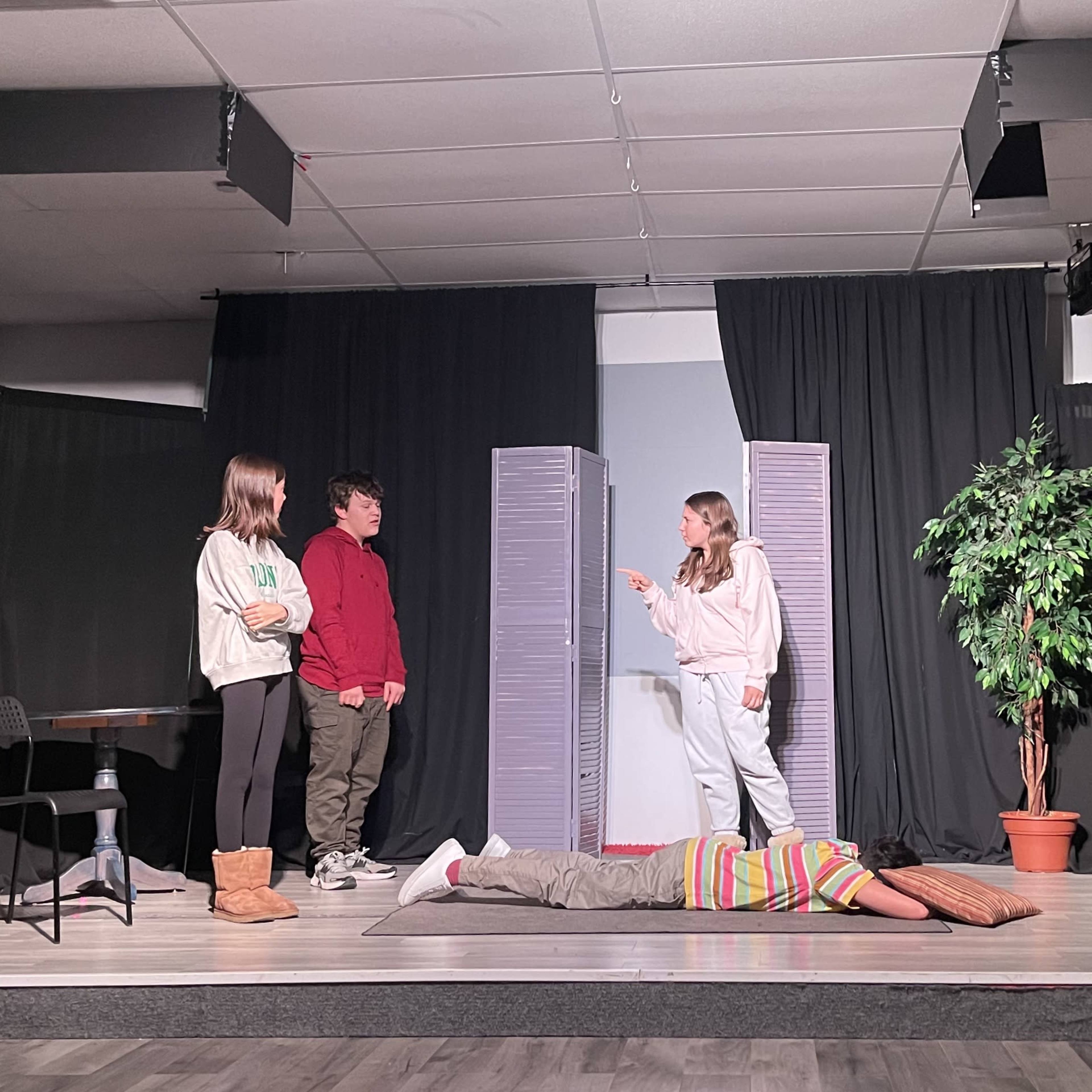A group of five actors is performing on a stage, with two standing, one lying on the floor, and two seated on a couch, amidst simple set pieces including curtains and a potted plant.