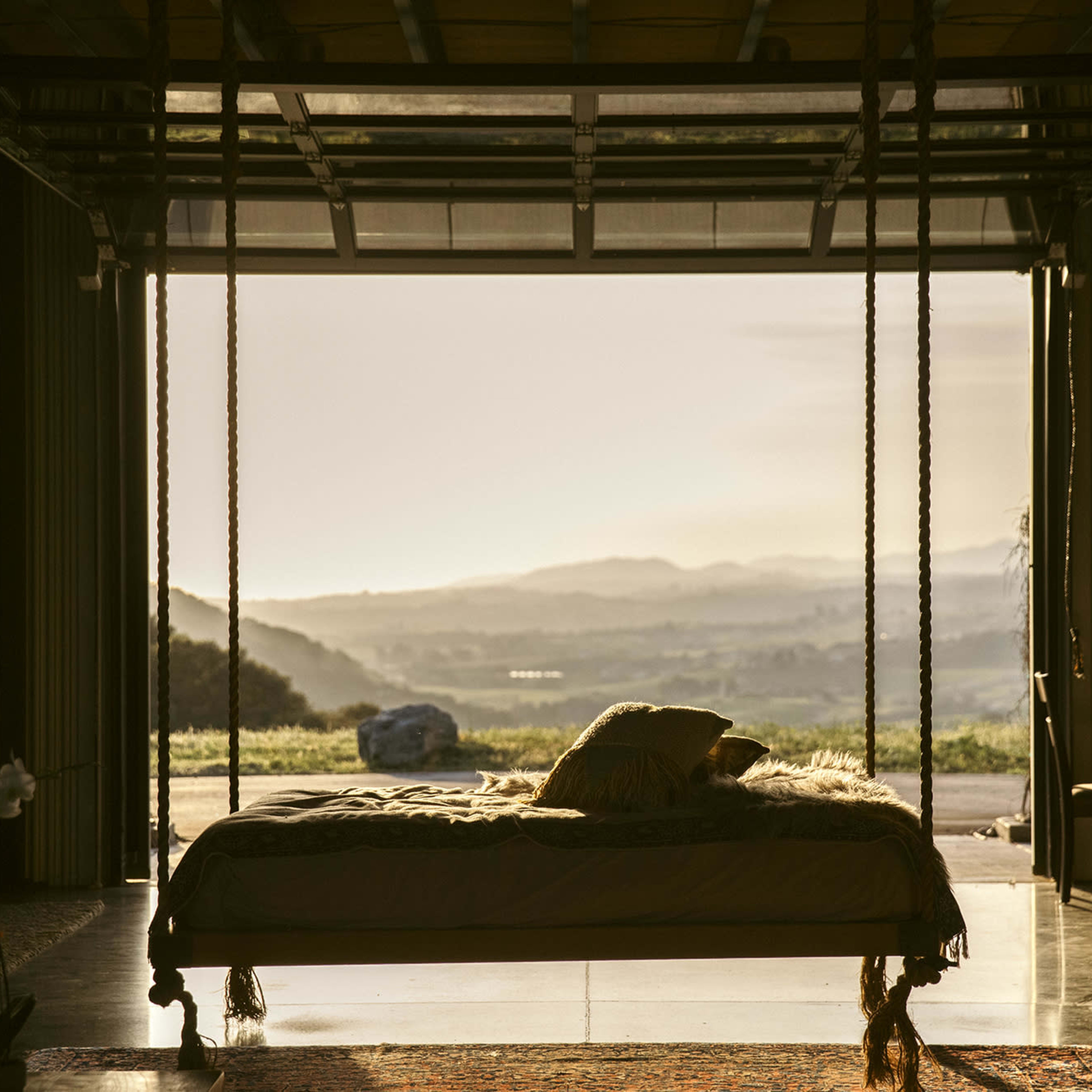 A bed with soft bedding is suspended from the ceiling in an open garage-like space that looks out onto a distant landscape at sunset.