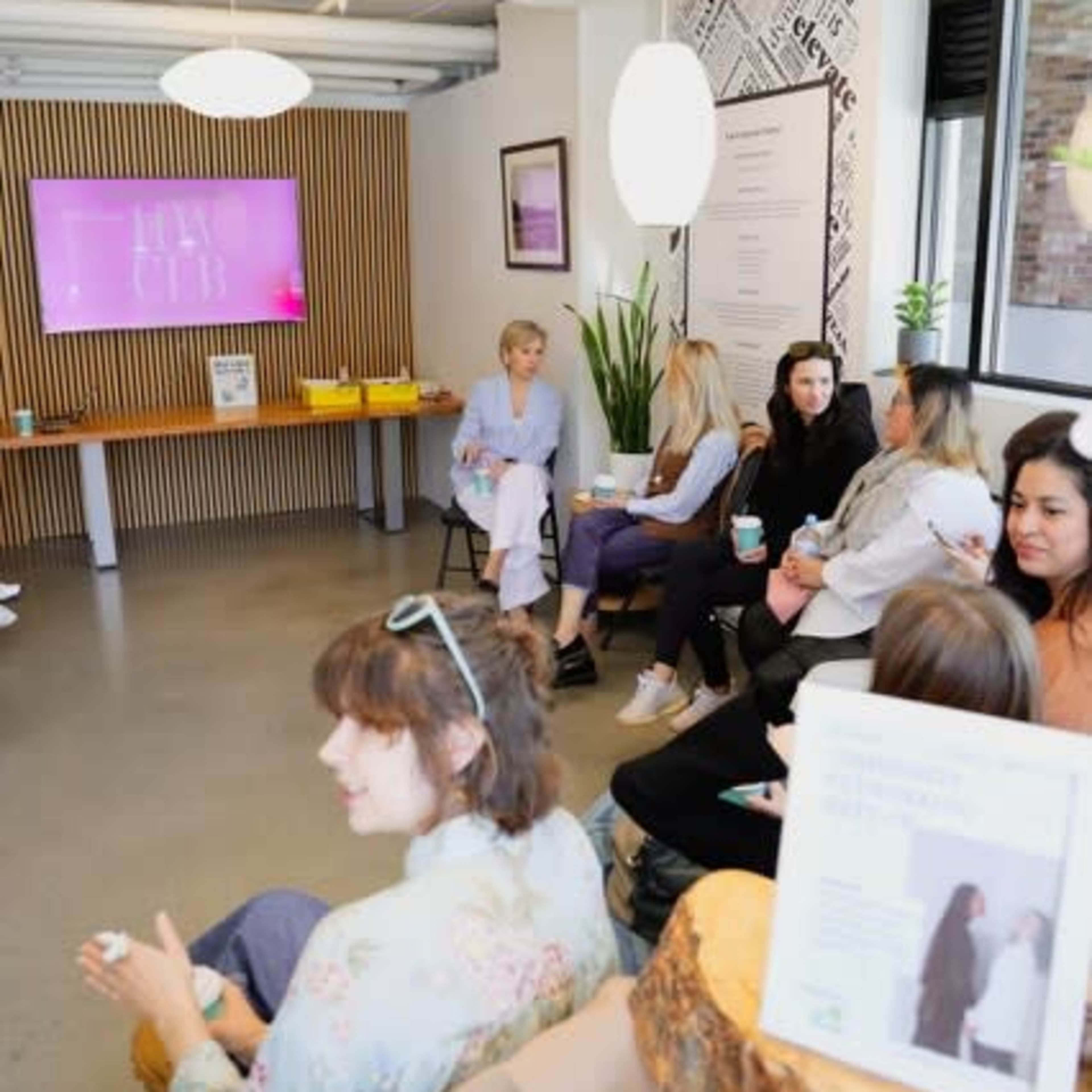 A group of people is seated in a modern, well-lit room with a presentation screen displaying "HUB," while a few individuals converse and hold drinks.