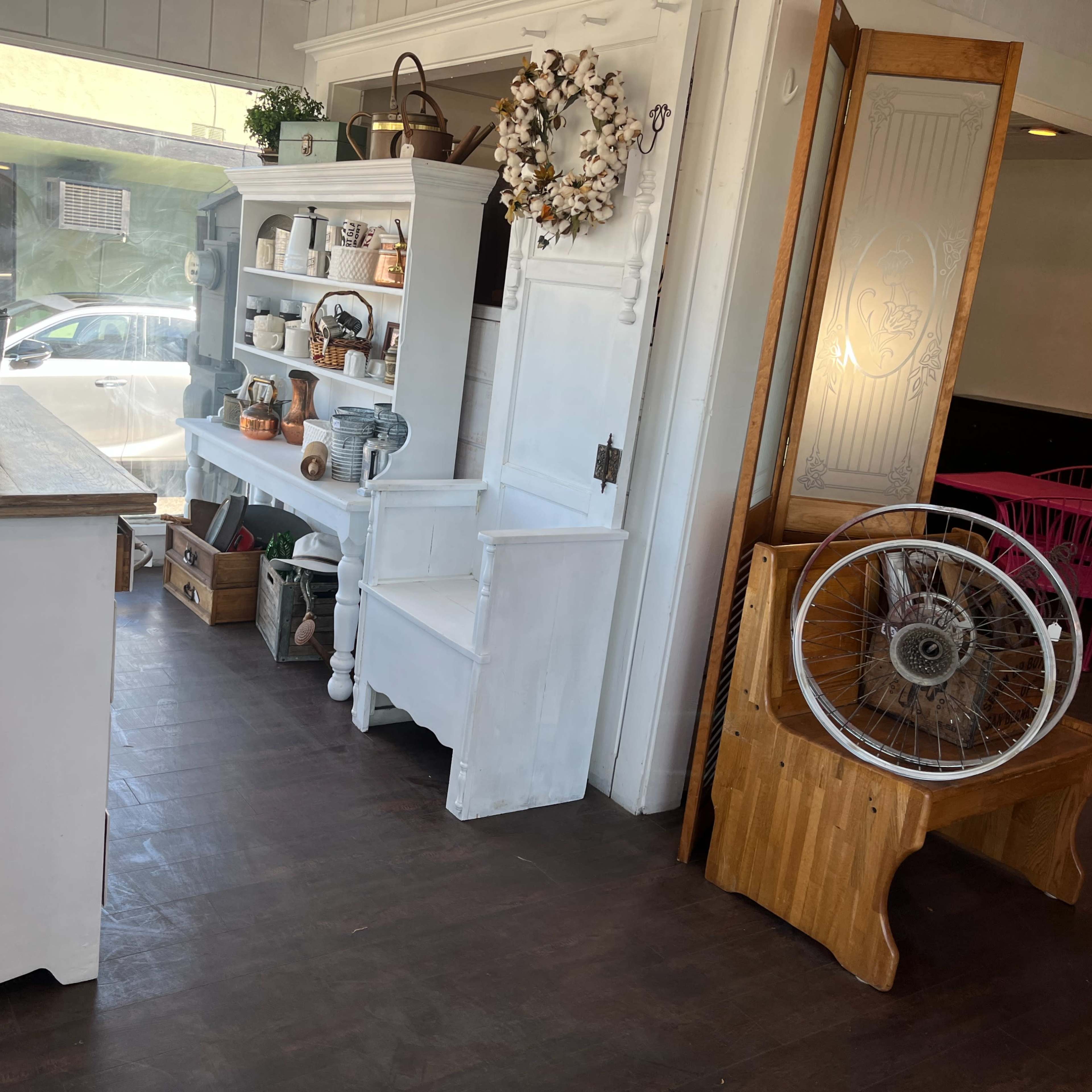 A cozy cafe interior with a wooden table, a display shelf of kitchen items, and a decorative wheel mounted on a wooden bench.